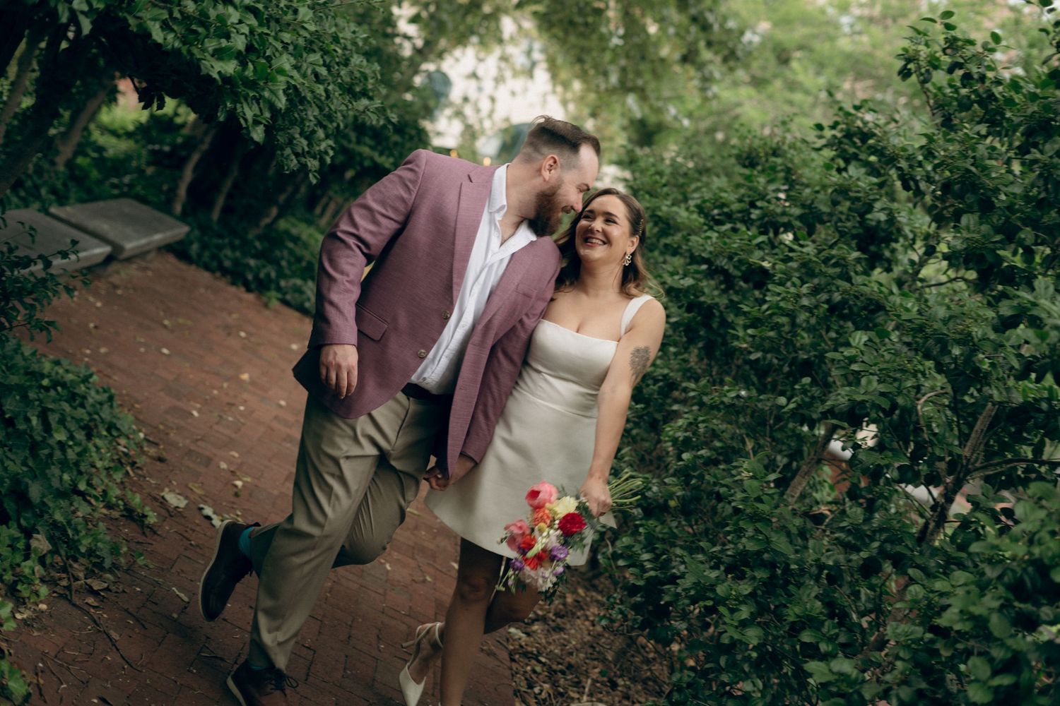 A couple in wedding attire walks hand in hand along a wooded path surrounded by lush green foliage.