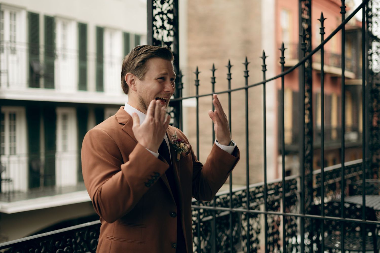 Someone in a brown coat gestures expressively while standing by an ornate wrought iron fence on a city balcony.