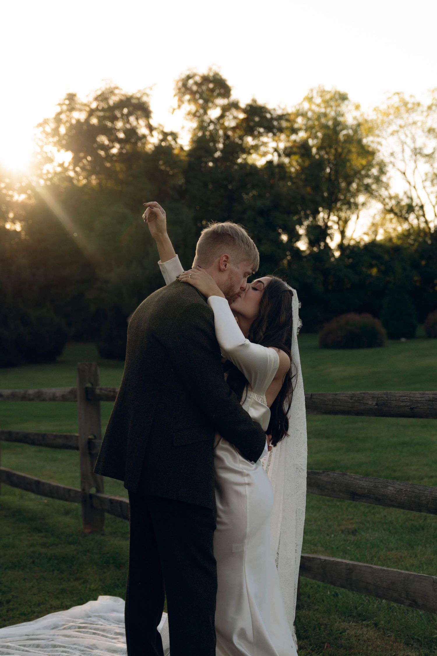 Romantic sunset embrace between newlyweds by countryside fence.