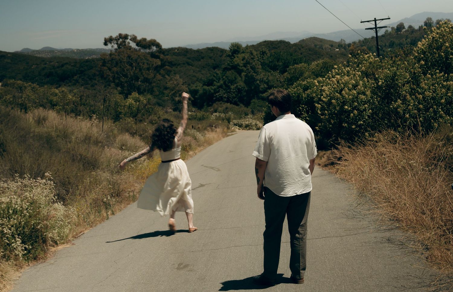 Two people walk together on a hilly rural road surrounded by dry brush and utility poles on a sunny day.