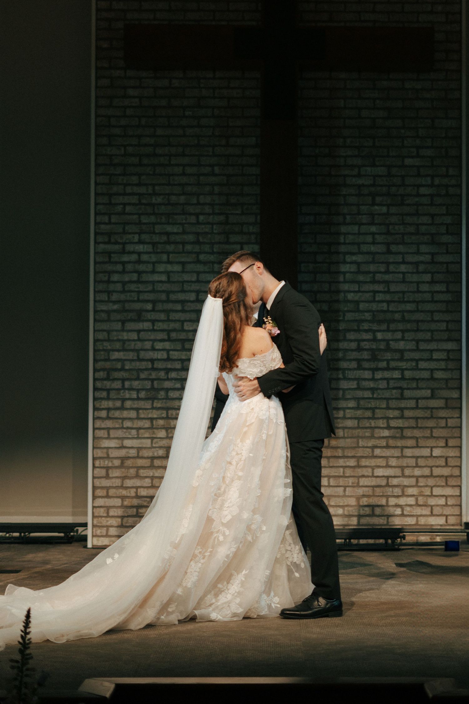 Wedding couple in romantic embrace against brick wall during artistic dark photo shoot with flowing white lace dress and veil.