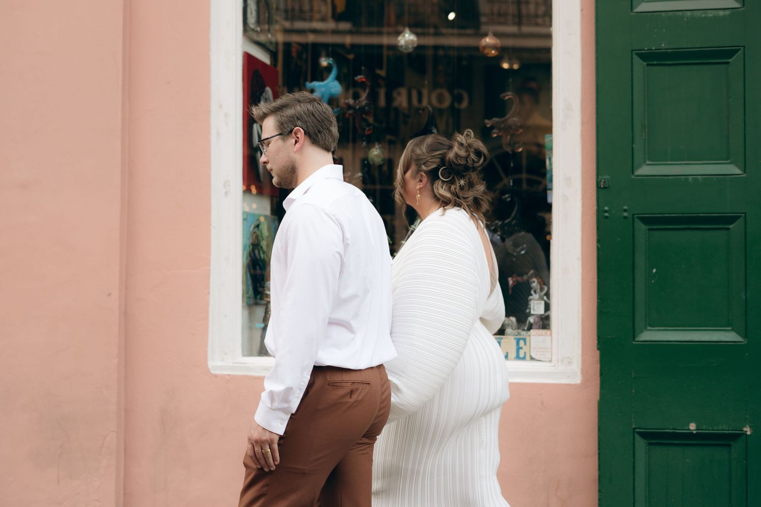 A couple in white outfits stands back-to-back outside a storefront with a green door.