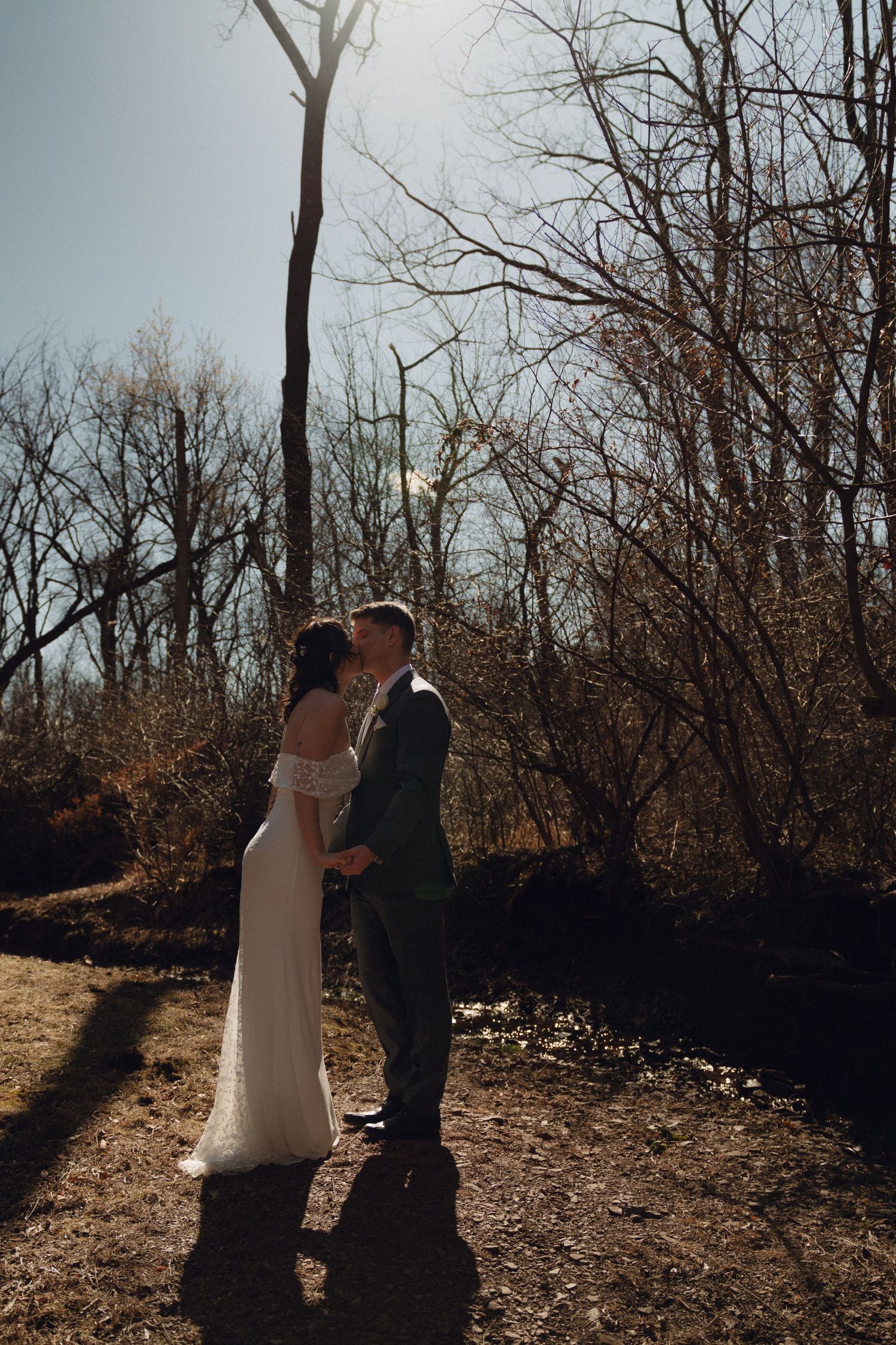 Couple in wedding attire kissing in silhouette against golden sunlight filtering through bare winter trees.