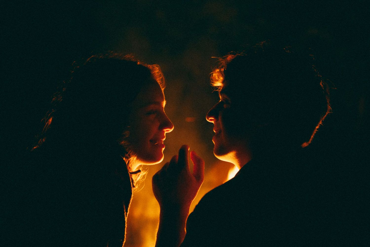 Two people facing each other illuminated by warm golden firelight against a dark background.