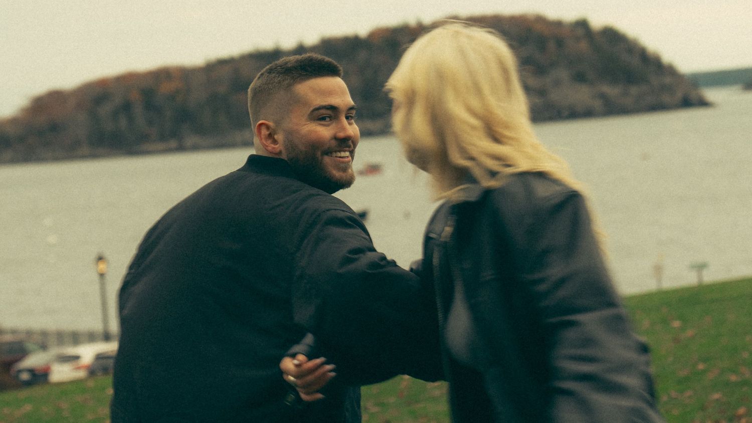 A couple shares an intimate moment while standing on grass overlooking a waterfront landscape. Couple in Bar Harbor Maine
