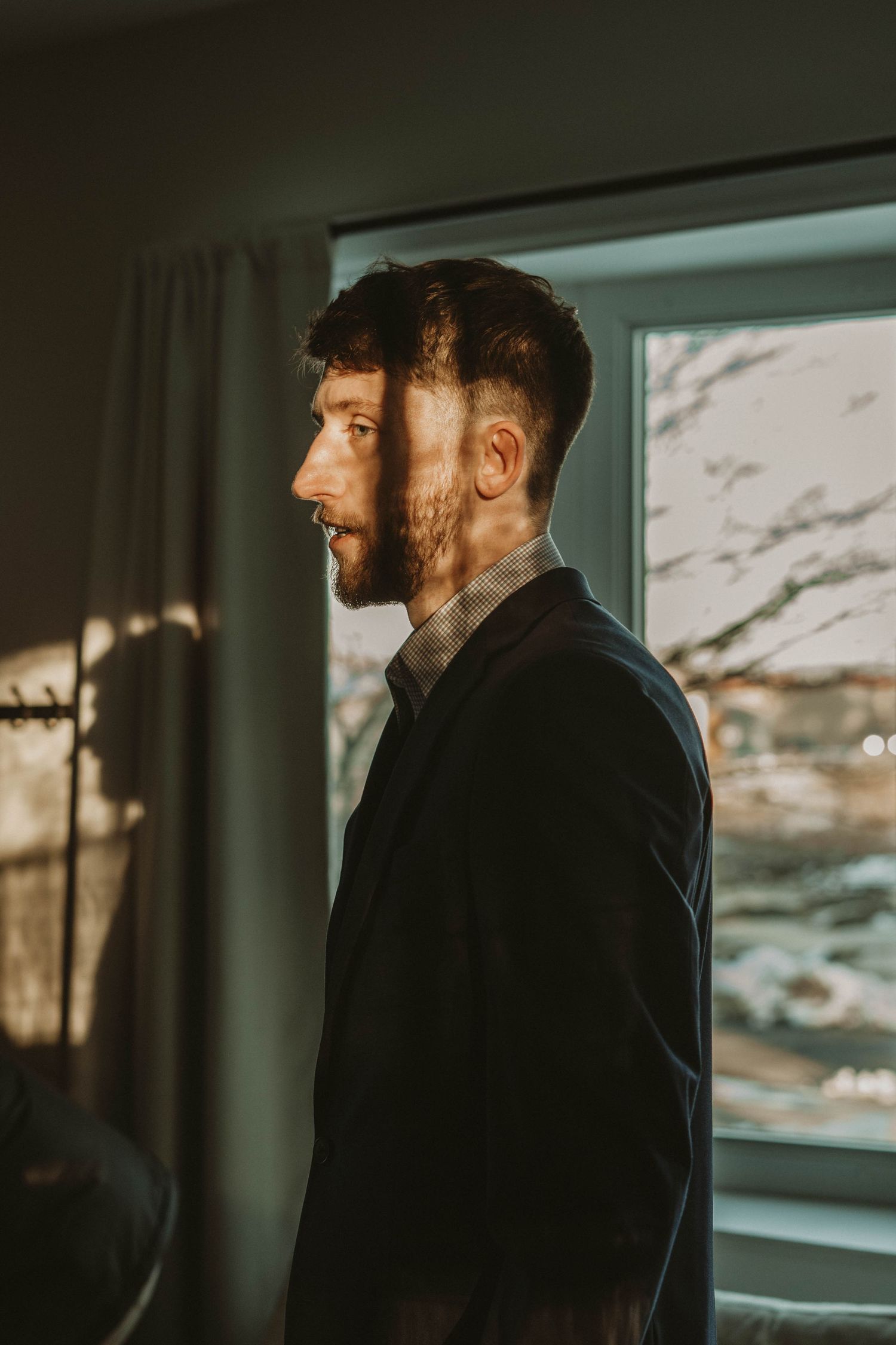 A person in a dark suit stands in profile by a window with natural light streaming in.