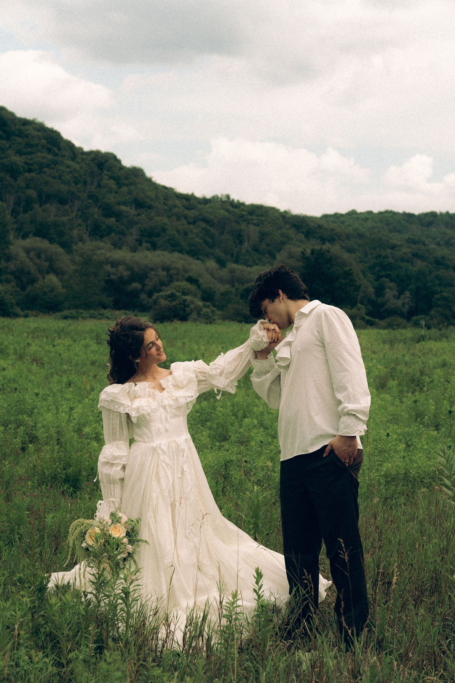 Bride in white gown sits in green field as groom kisses her hand, with lush forested hills in background.