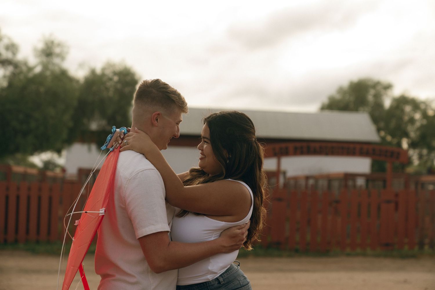 Young couple embracing outdoors near a red fence, sharing a romantic moment on a cloudy day.