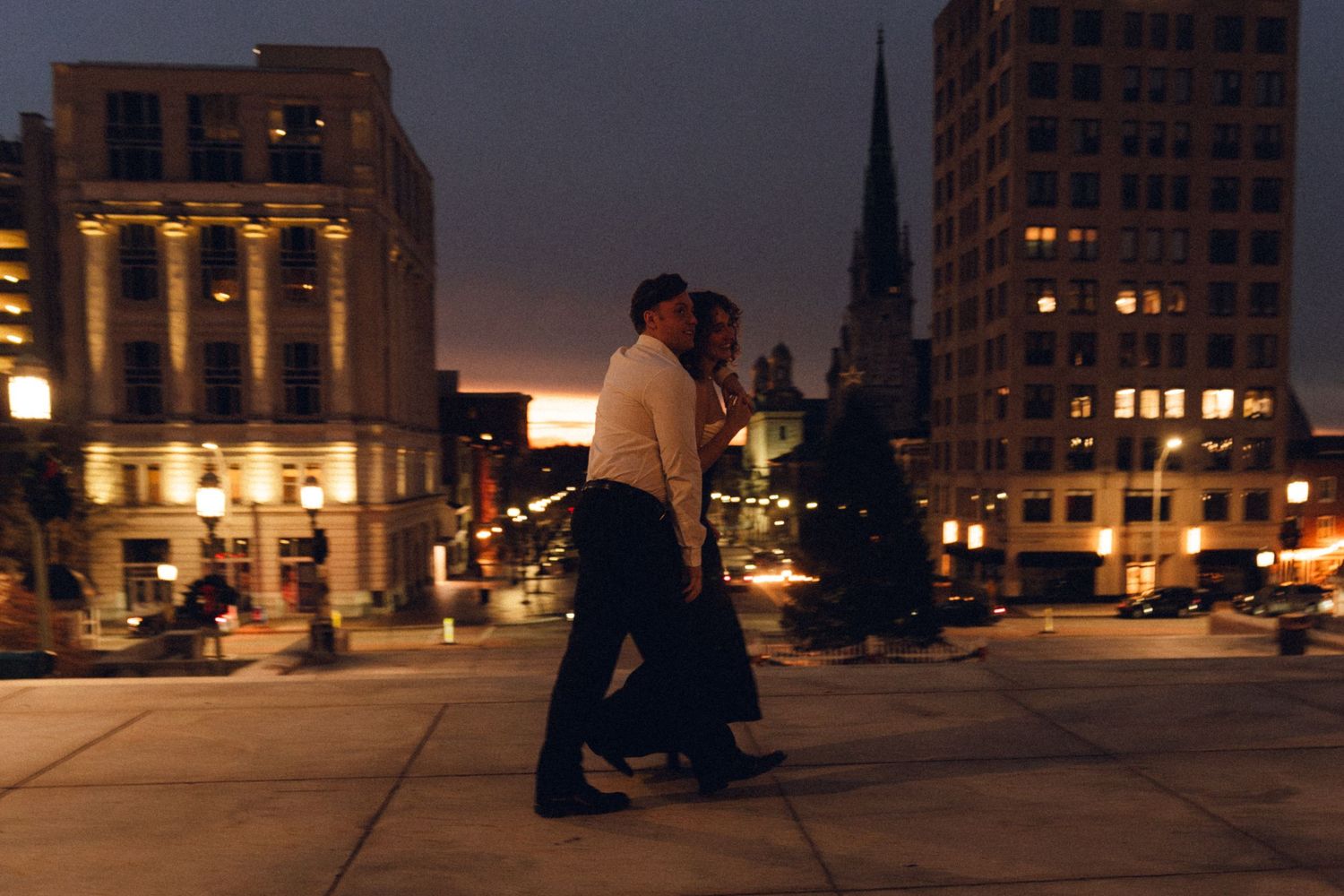 Couple walking together on a rooftop at night with city buildings and glowing lights in the background.