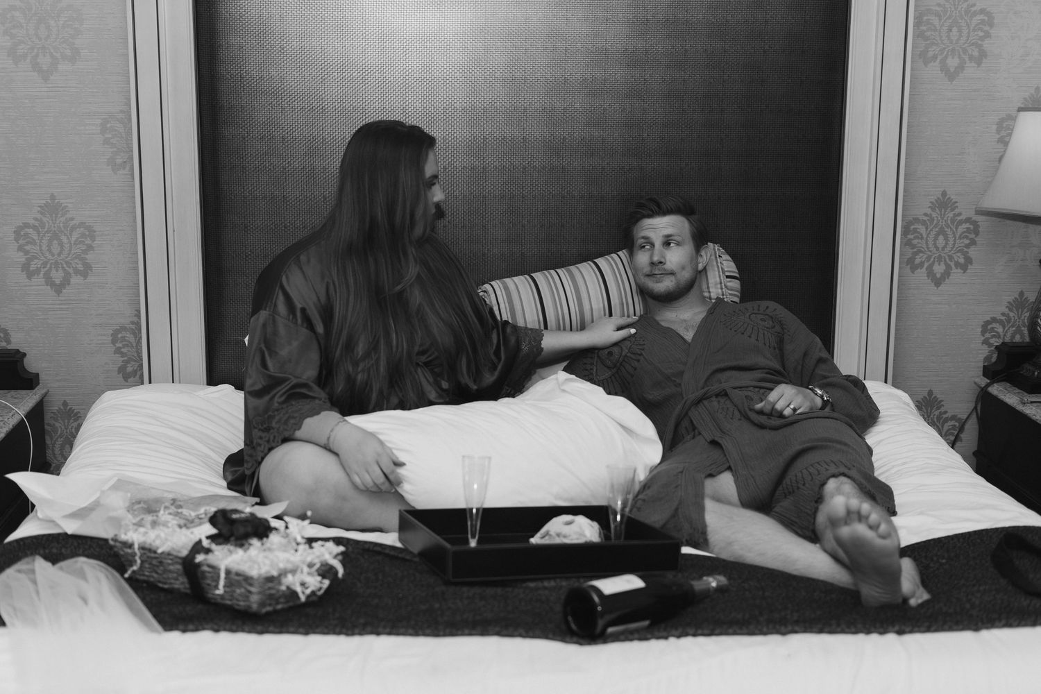 Two people relax on a bed while having a casual conversation in a simple bedroom setting with vintage decor.