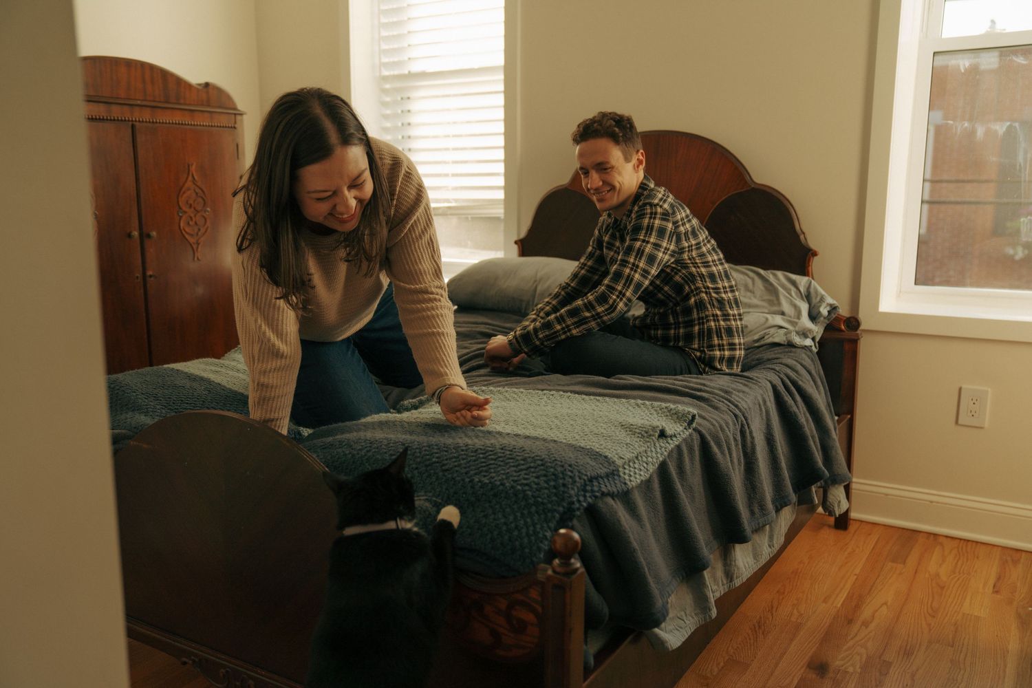 Two people sit on a blue bed in a cozy bedroom with hardwood floors and warm lighting.