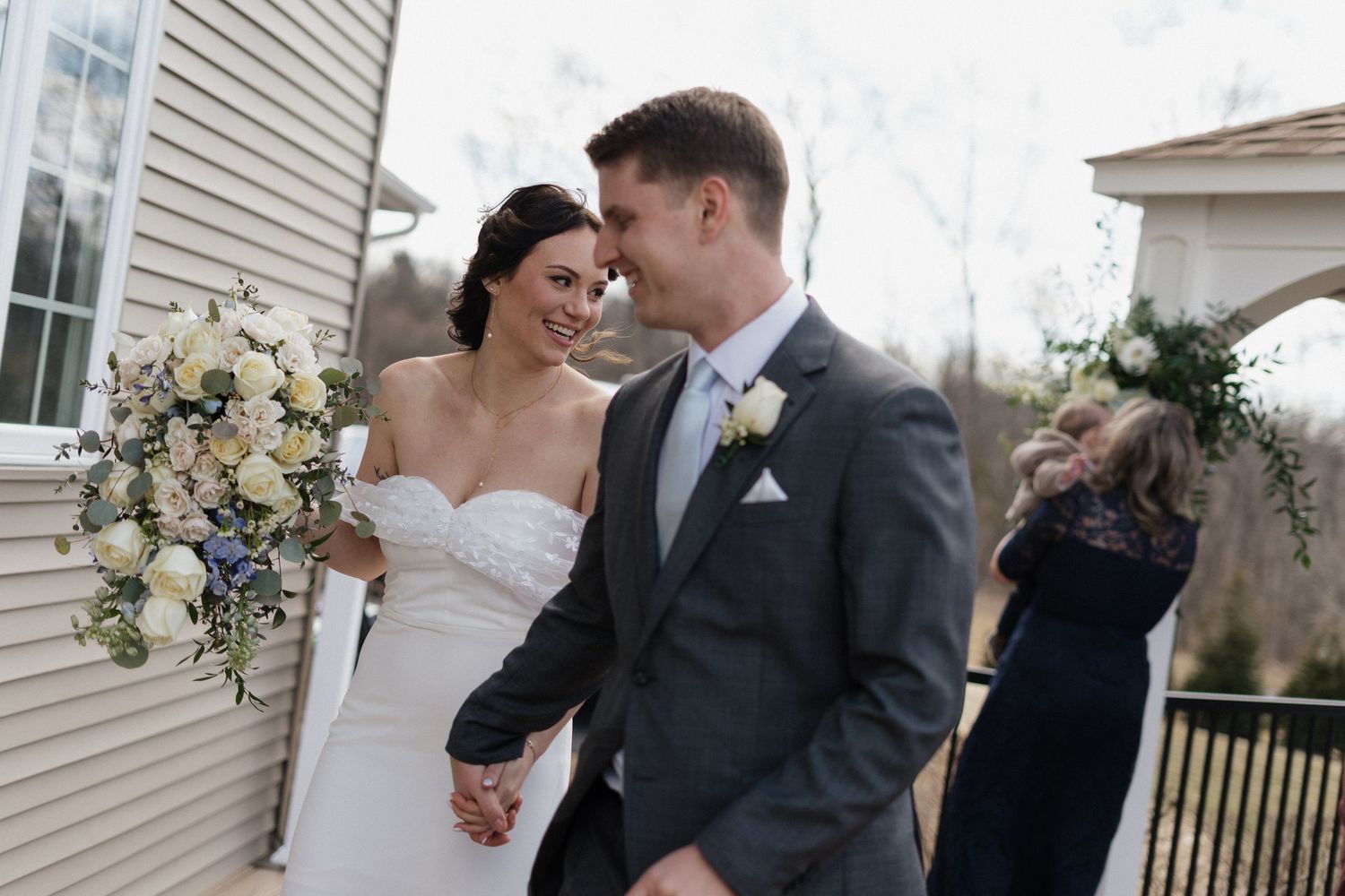 A bride in white dress holds a bouquet while sharing an intimate moment with her groom outdoors.