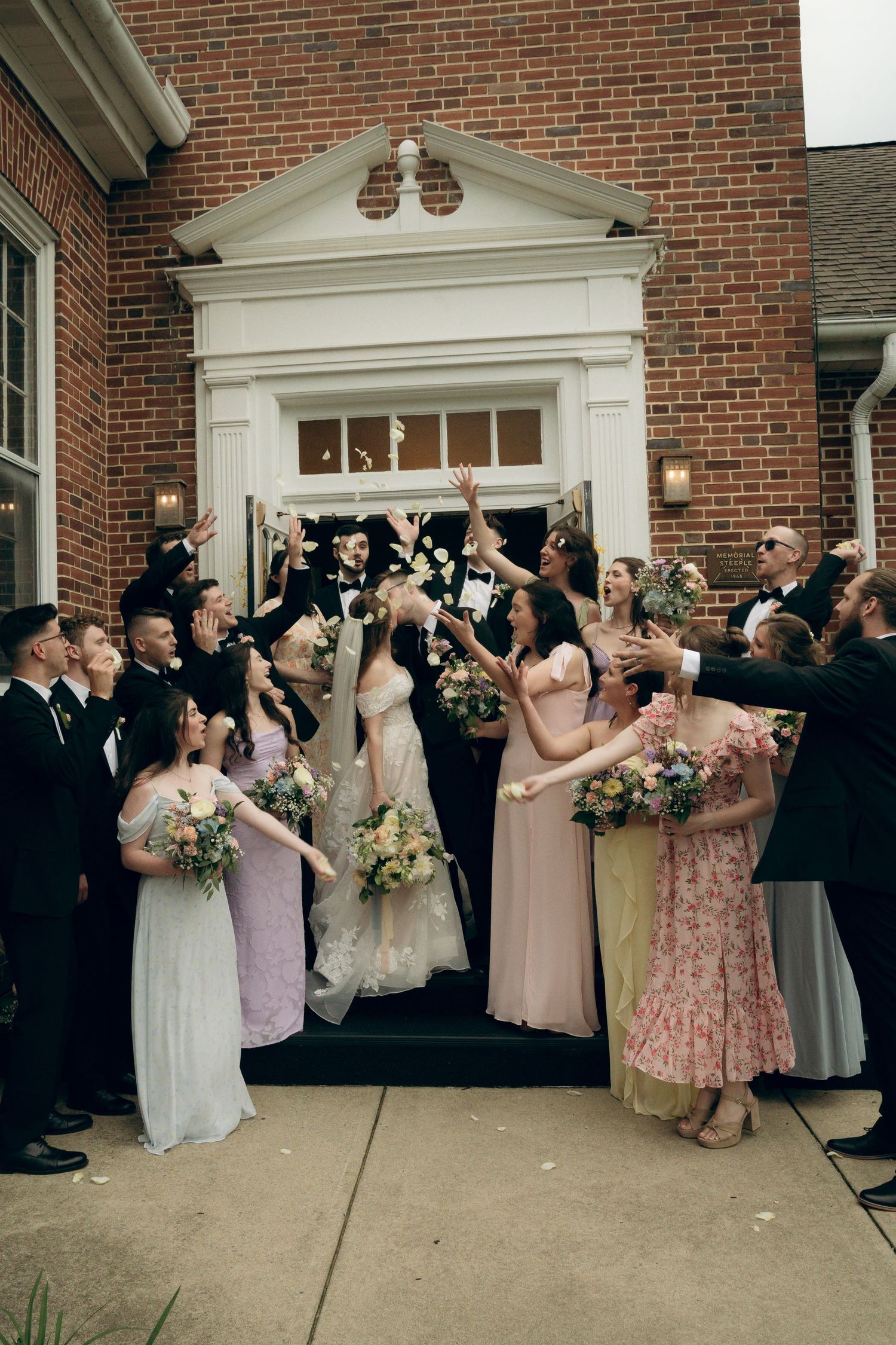 Wedding party celebrates outside brick church entrance by tossing flower petals in the air.