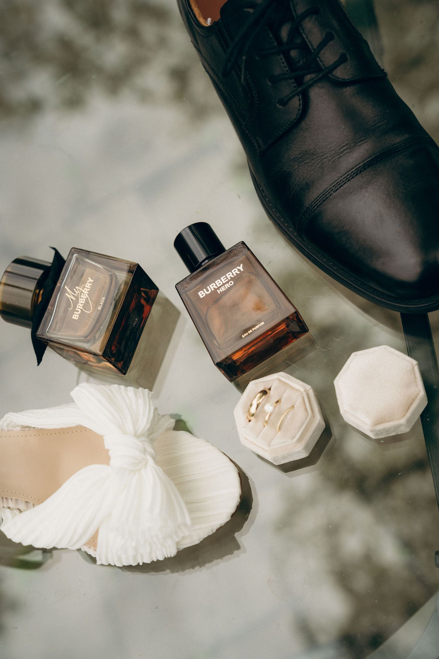 Wedding accessories including formal black shoes and perfume bottles arranged on reflective surface.