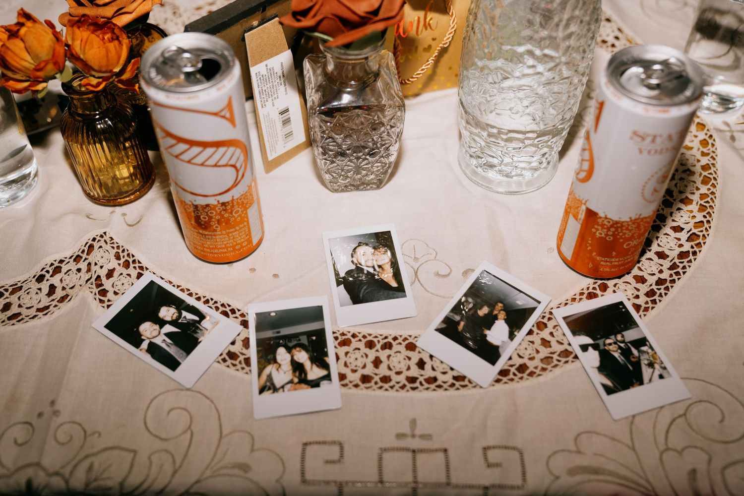 Wedding table decor with polaroid photos, orange roses, and drink cans scattered on a lace tablecloth.