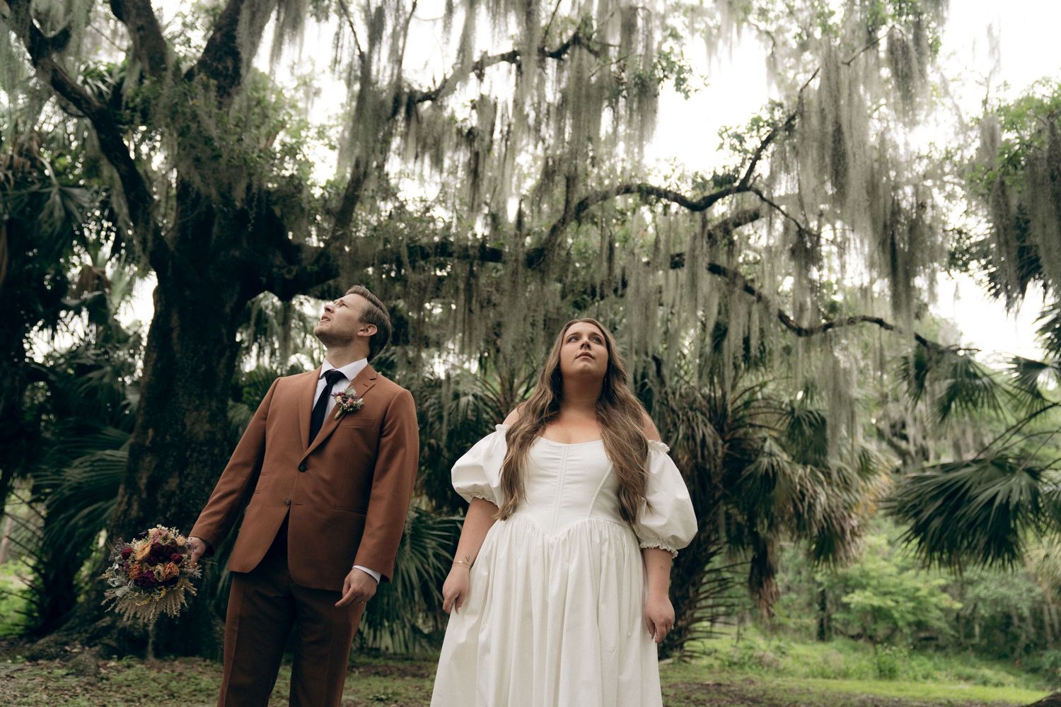 Couple in wedding attire poses beneath Spanish moss draped trees in a romantic outdoor setting.