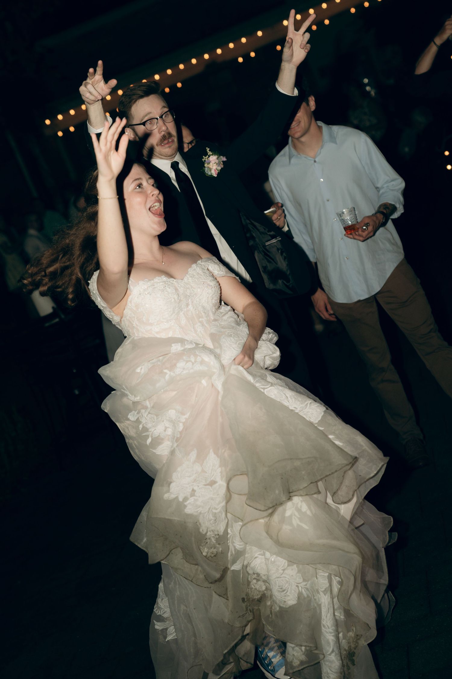 Wedding celebrants dance enthusiastically on a dark dance floor under string lights, with the bride wearing a layered lace gown.