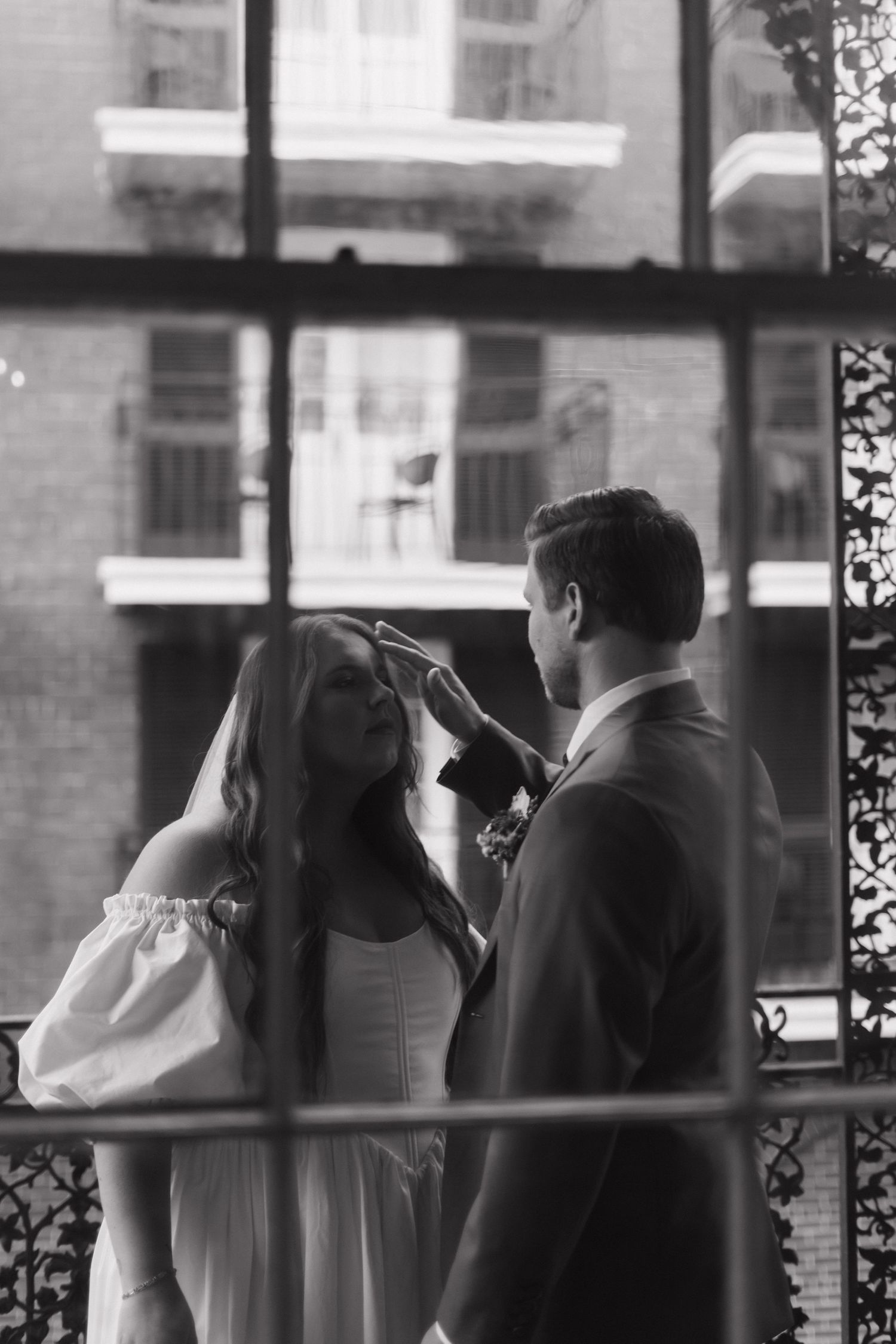 Black and white romantic moment captured through a window showing a tender interaction between a couple in formal attire.