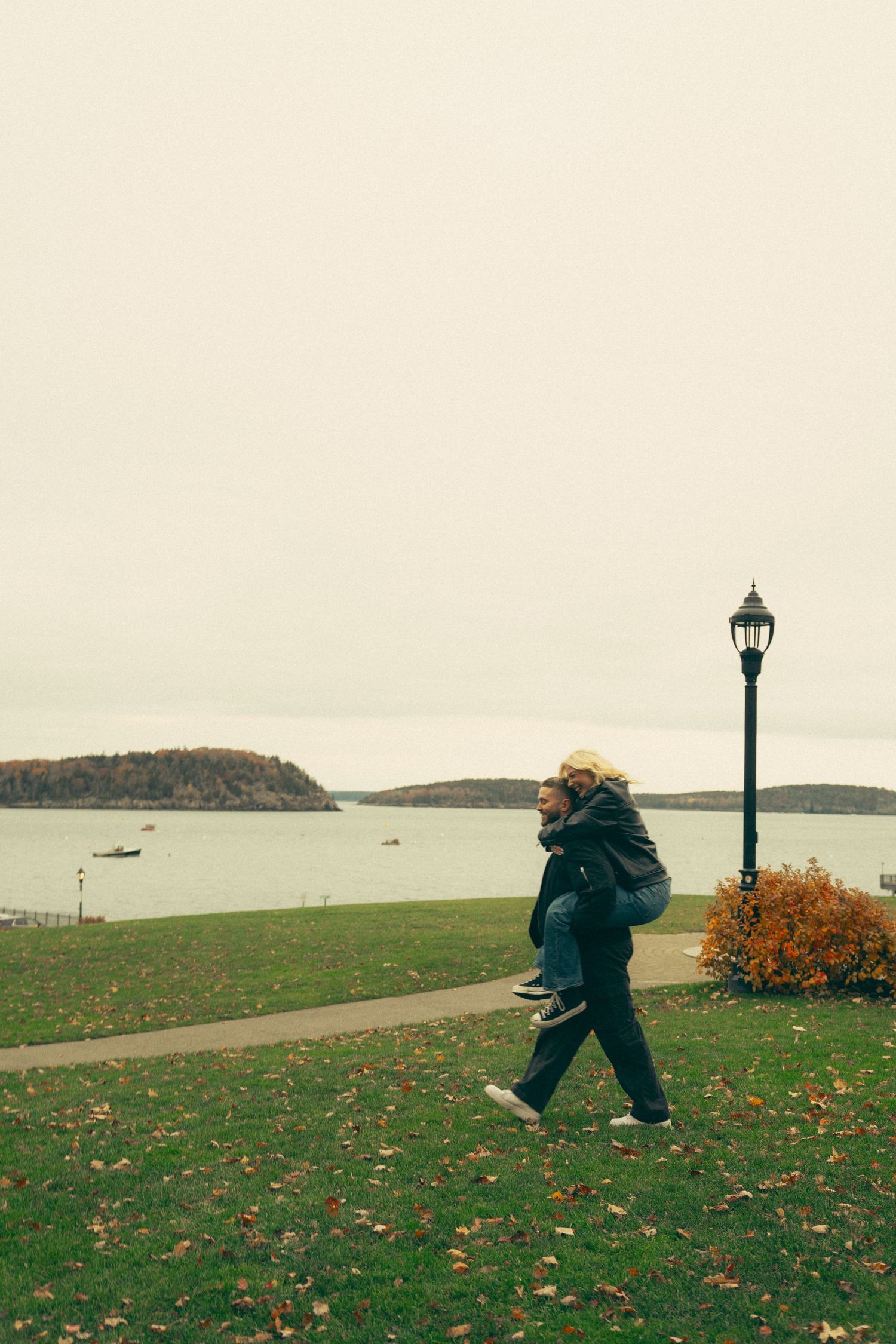 Person giving piggyback ride near waterfront park with lamp post on overcast autumn day.