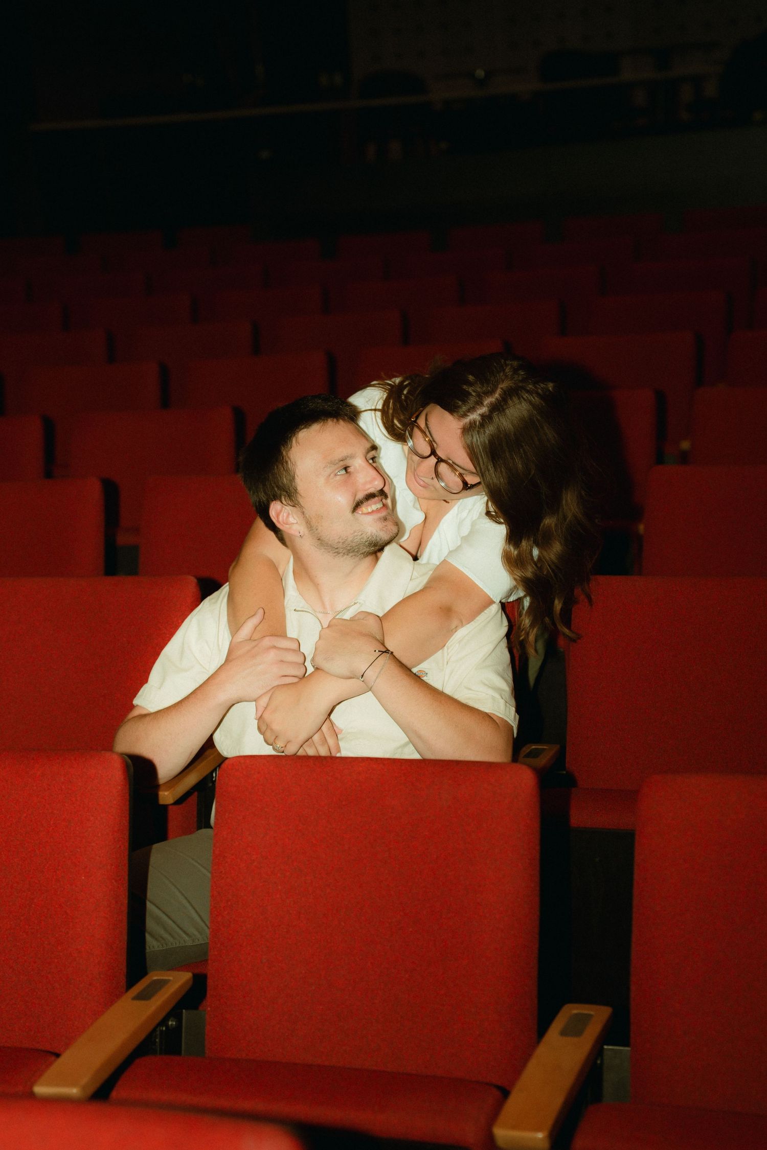 Two people share an intimate moment sitting close together on red theater seats.