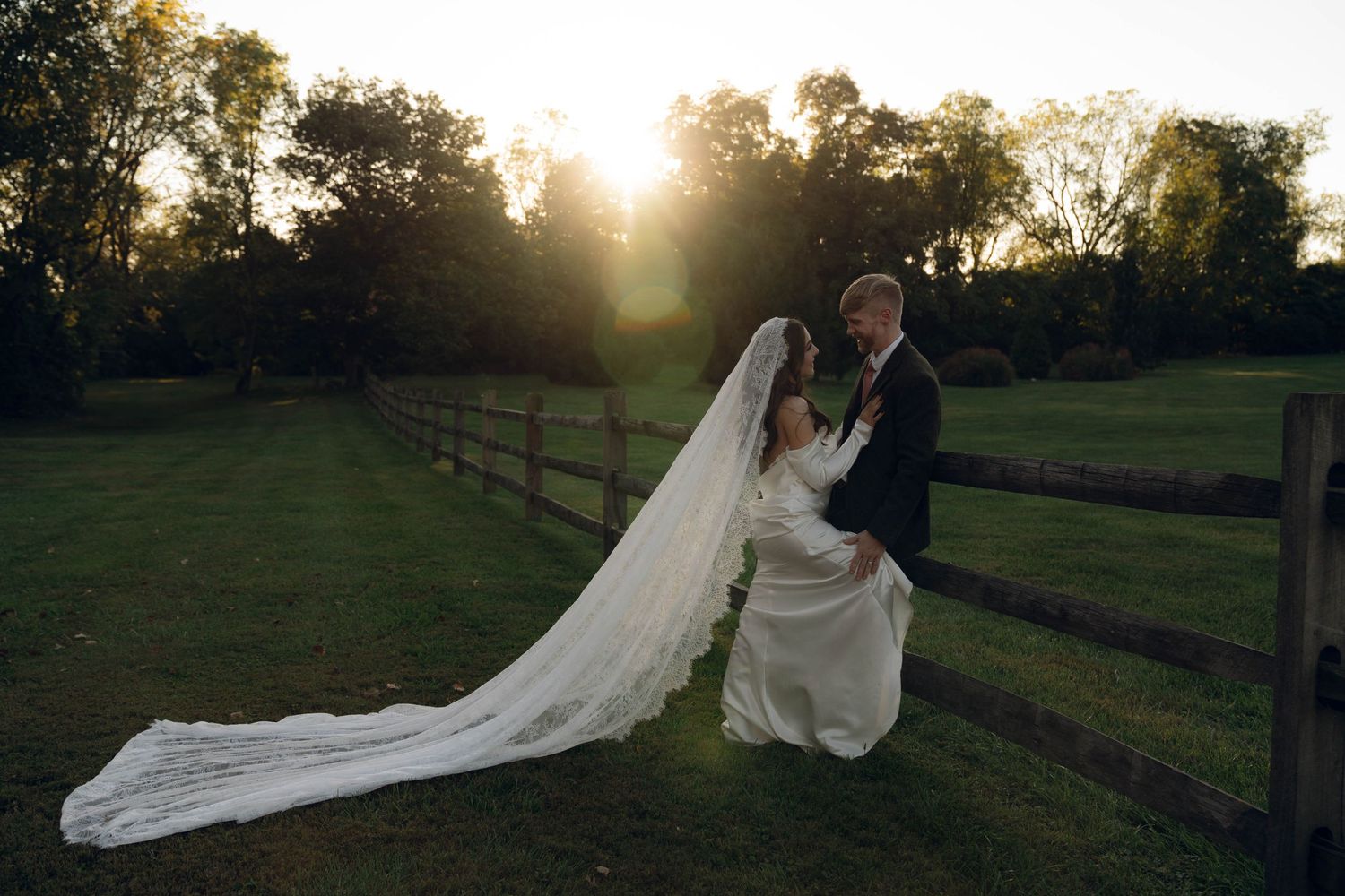 Sunset wedding portrait by rustic wooden fence with flowing bridal veil.
