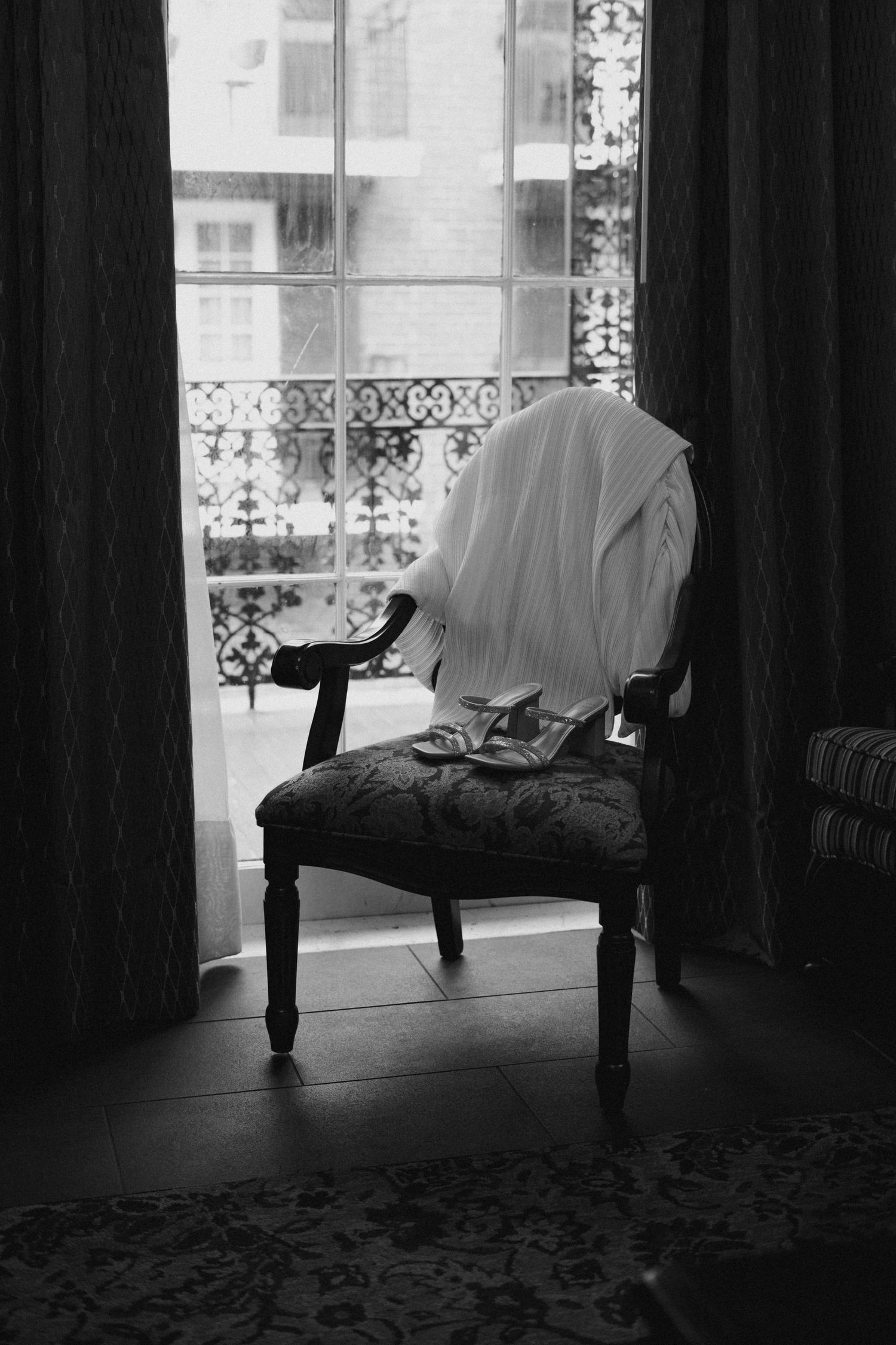 Black and white photo of a wedding veil draped over a vintage chair by a window.