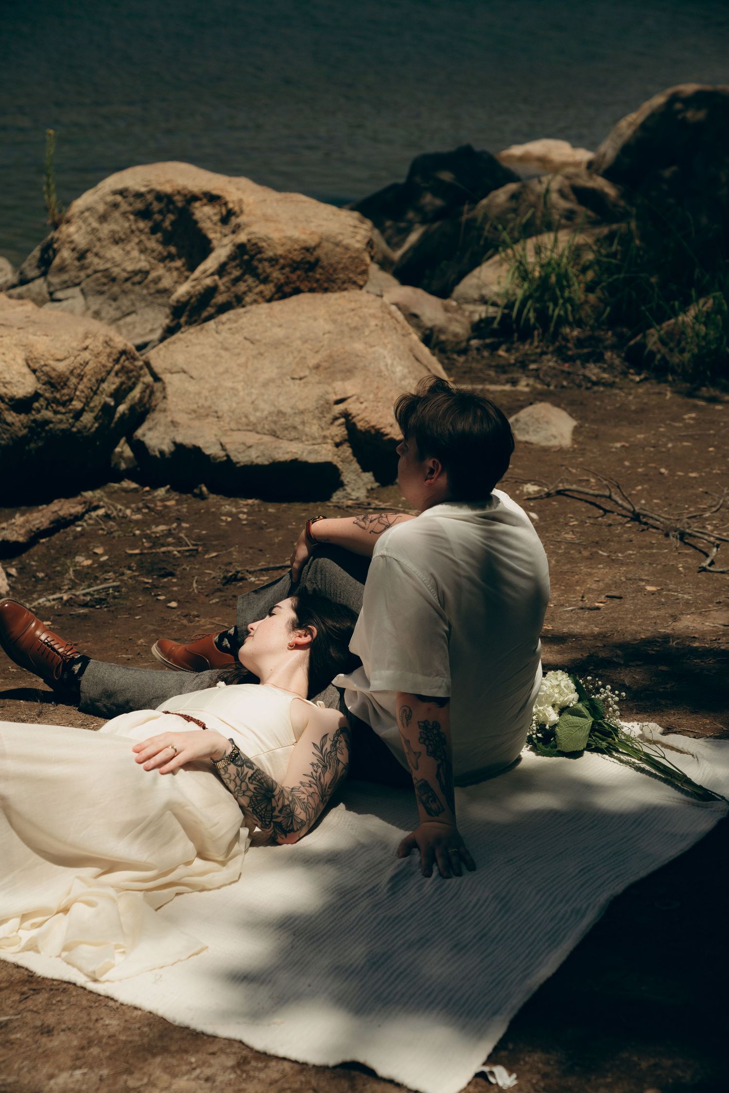 A couple relaxes on a white blanket near large boulders during an outdoor photoshoot.