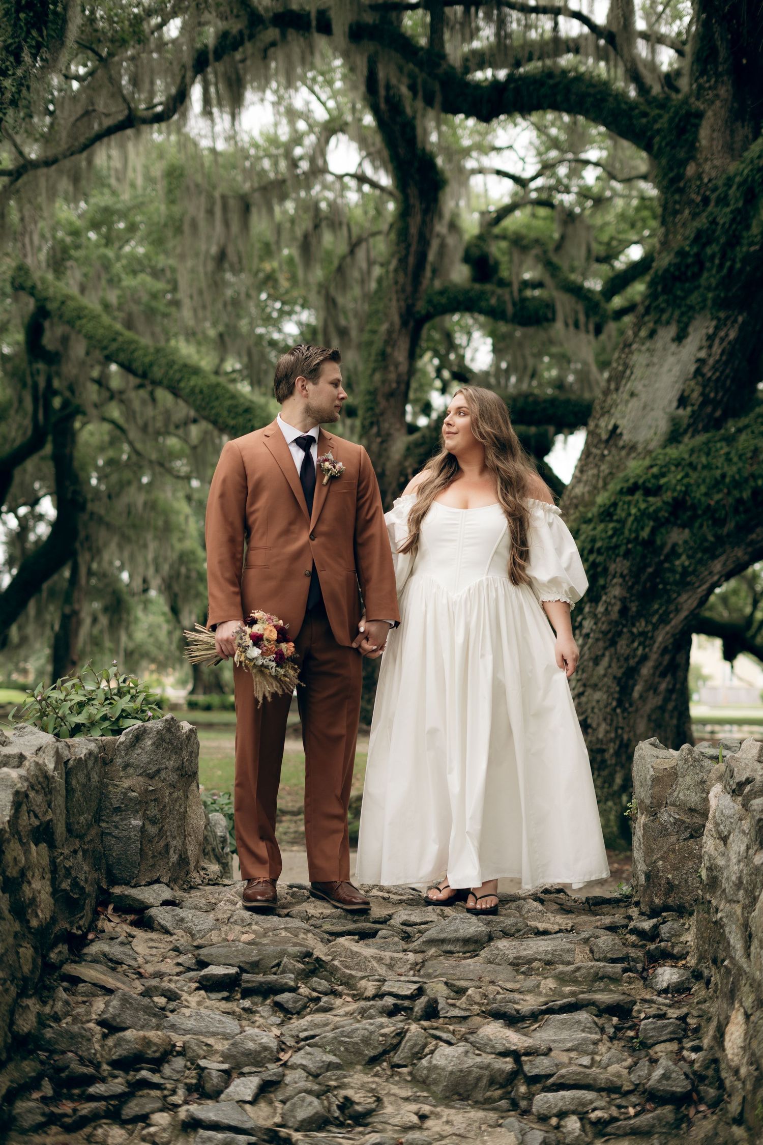 A couple in wedding attire stands on stone steps under moss-draped oak trees in a romantic outdoor setting.