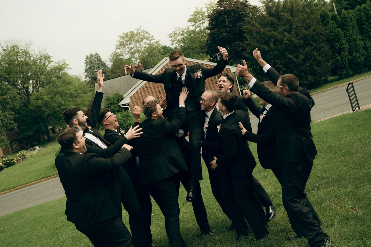 Wedding party dressed in black suits performs a synchronized jump pose on grassy lawn at outdoor celebration.