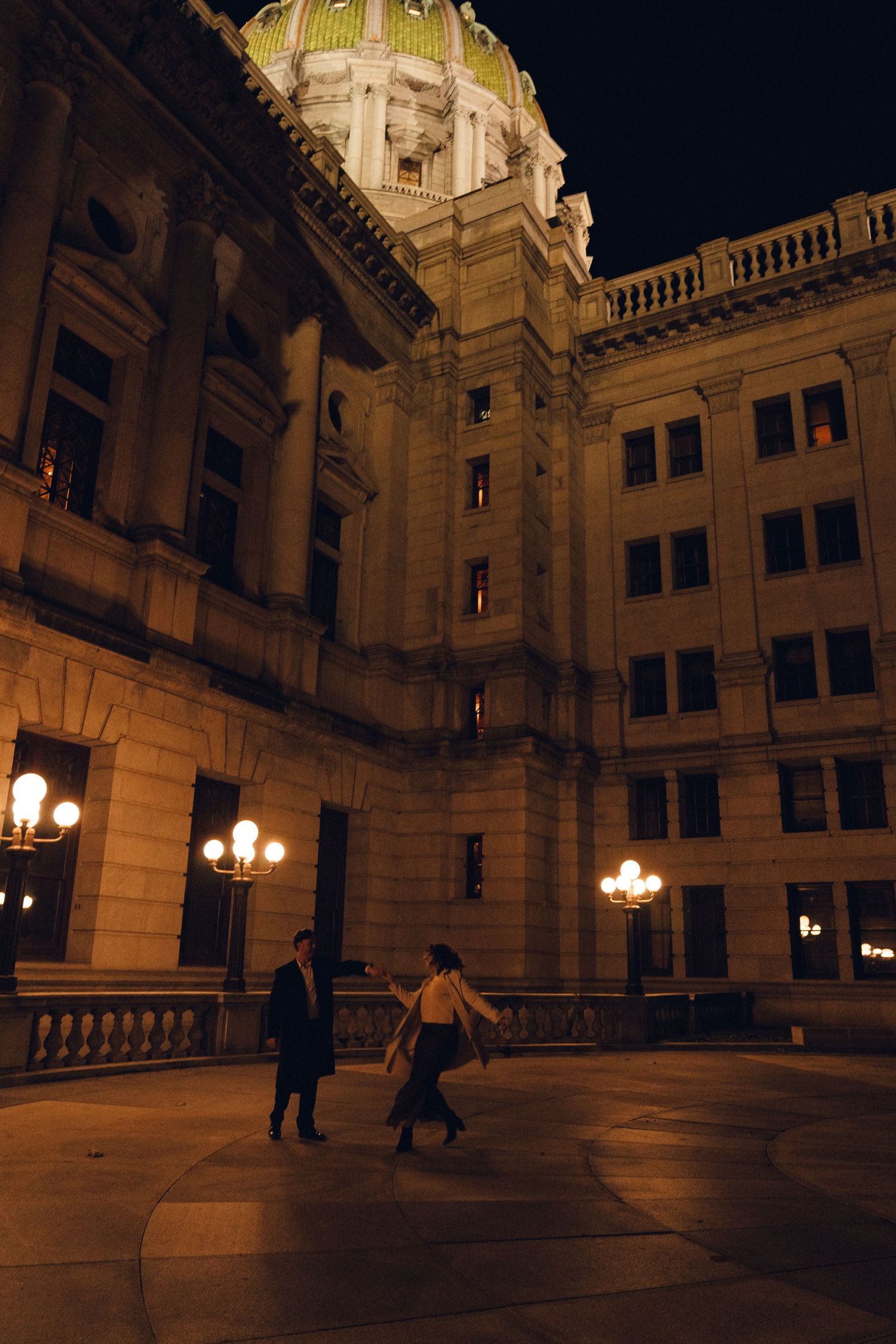 Couple walking at night outside illuminated neoclassical capitol building with green dome and ornate lamp posts.