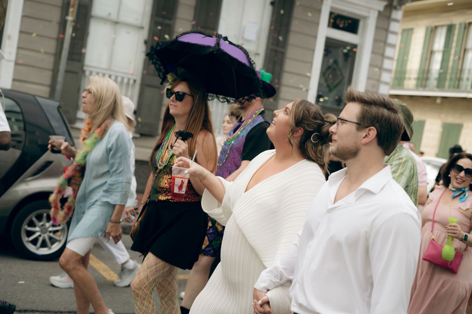 A lively street scene in New Orleans with partygoers holding drinks and wearing colorful costumes and parasols.