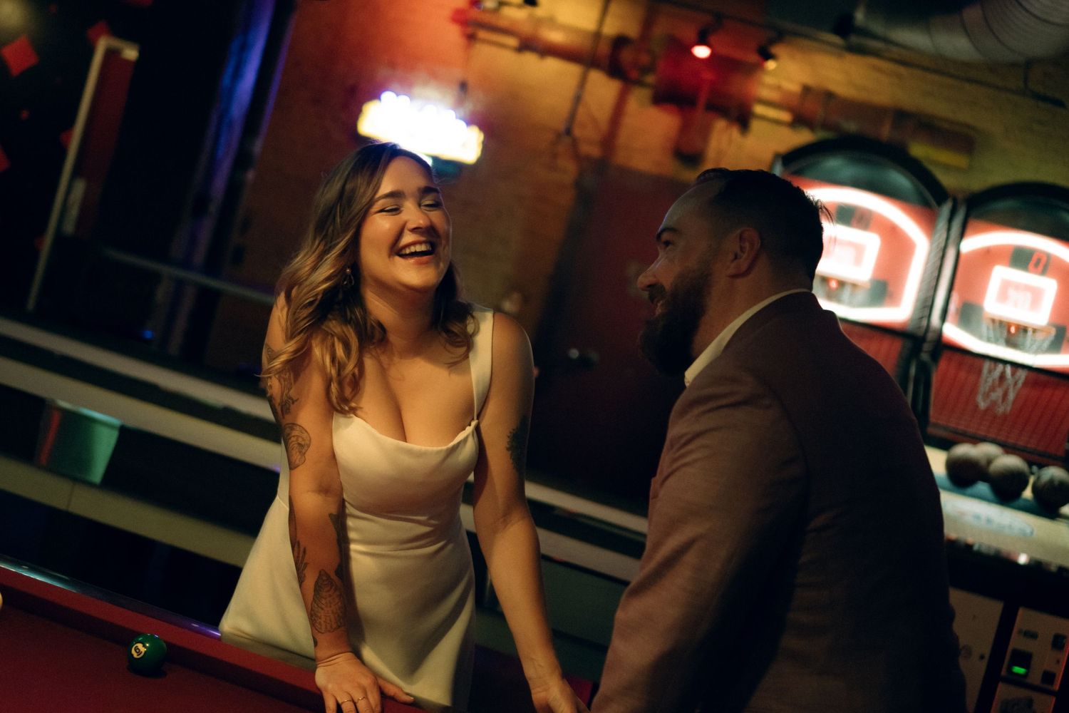 Two people laughing and talking while playing pool in a dimly lit bar with neon lights visible in background.