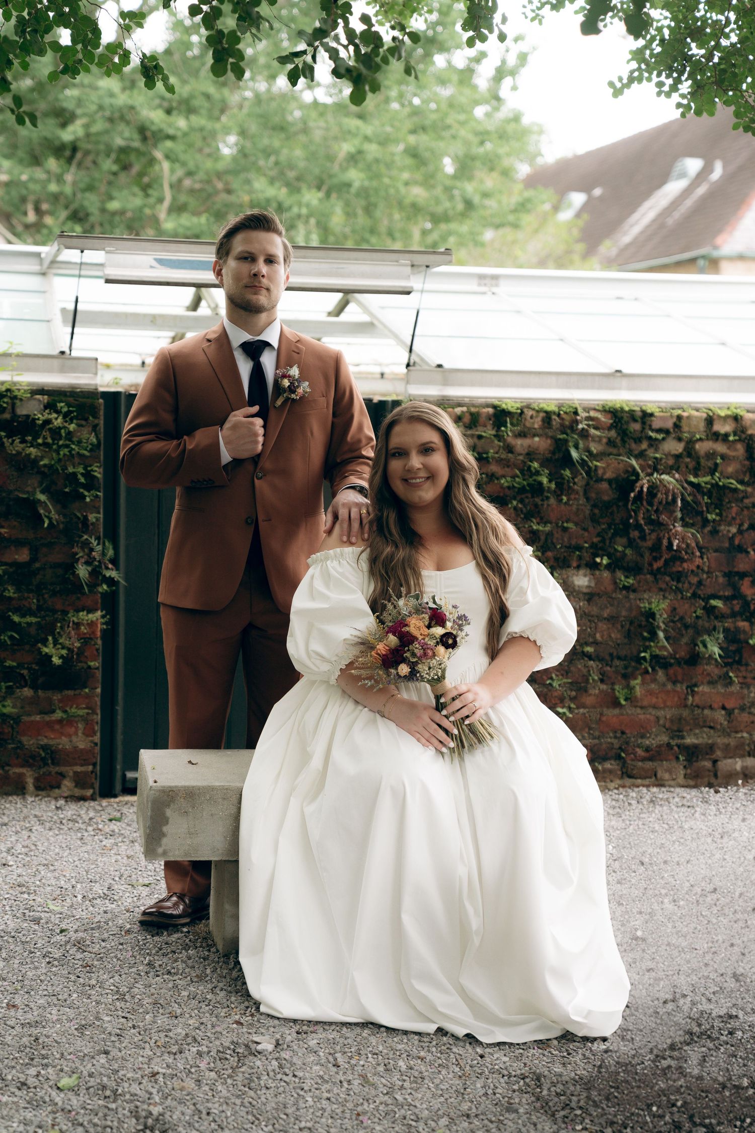 Newlywed couple shares a quiet moment while sitting together on wooden garden bench among brick walls.