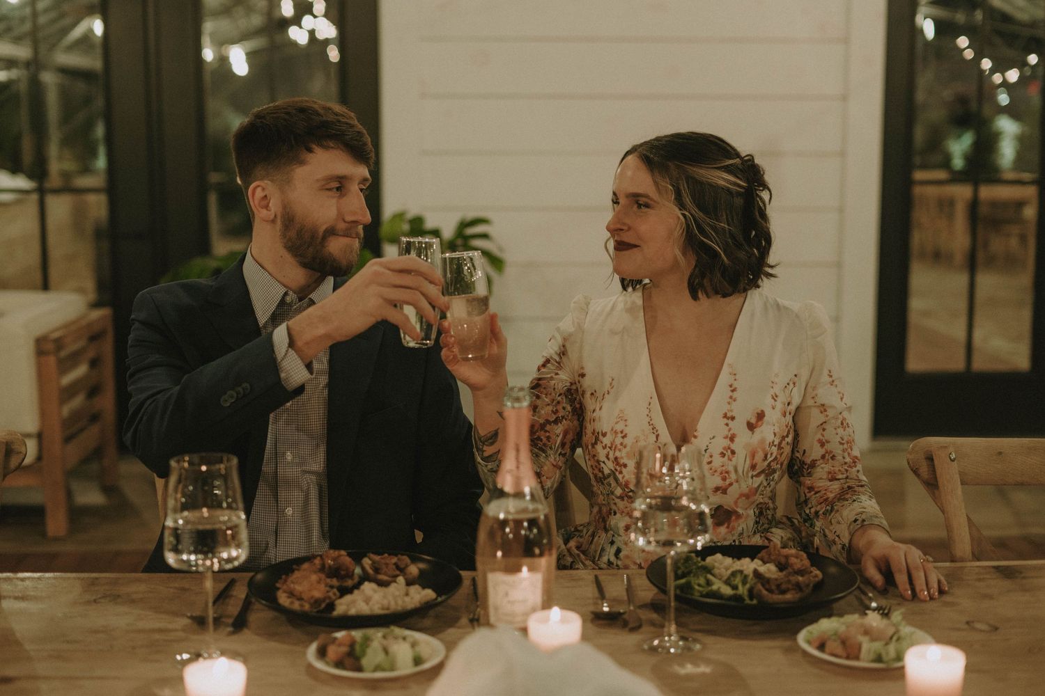 An intimate dinner scene with wine glasses and candlelight in a greenhouse setting.