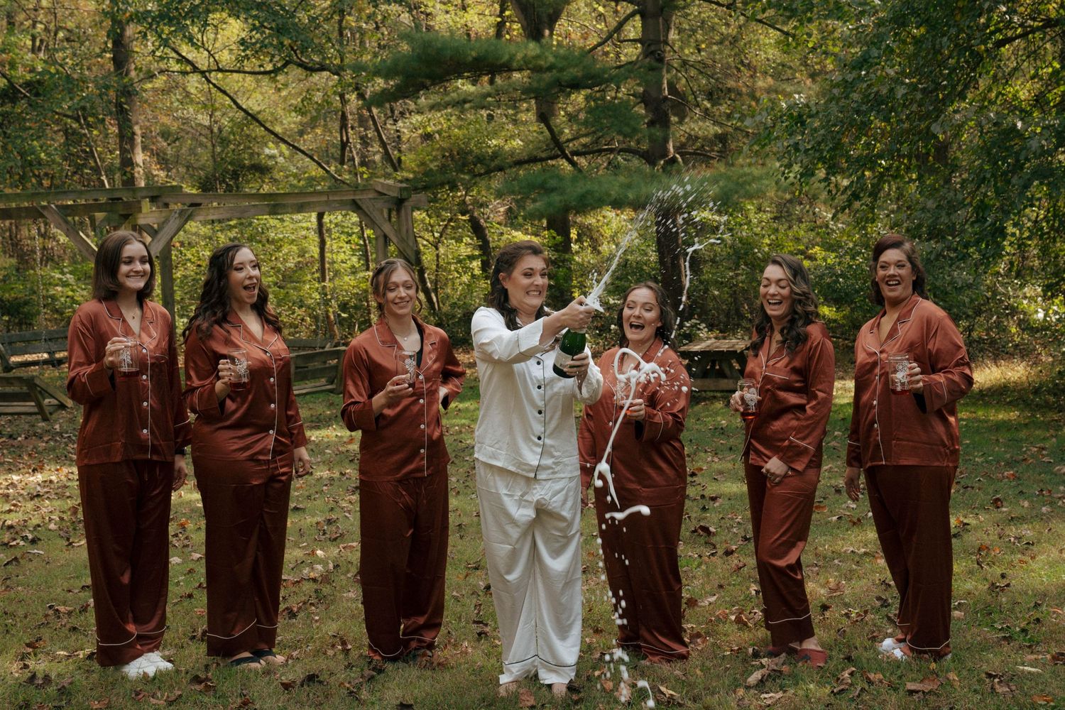 Wedding party in matching burgundy outfits poses with celebrant in white outdoors.
