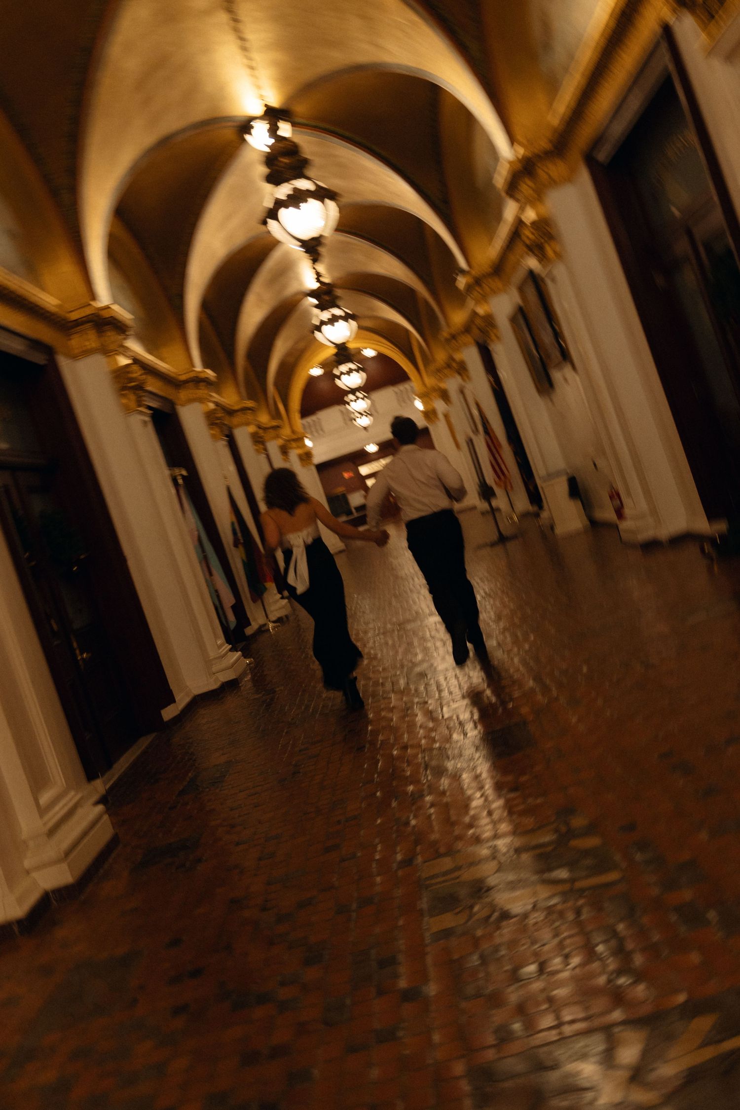 Dimly lit arched hallway with stone columns and cobblestone floor illuminated by warm ceiling lights creates a medieval atmosphere.