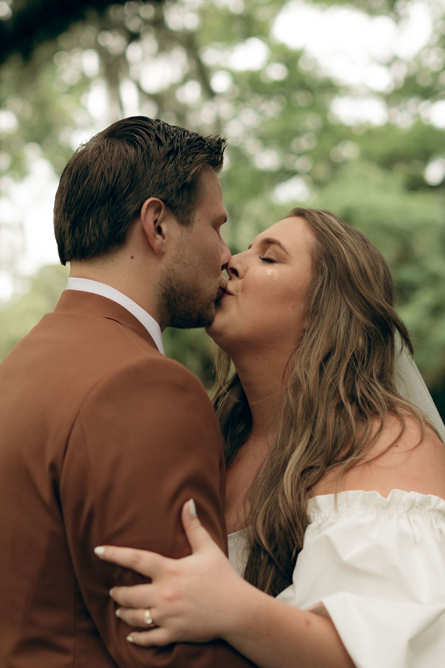 A couple shares a romantic kiss during their wedding day, with the bride in white and groom in brown against a soft green background.