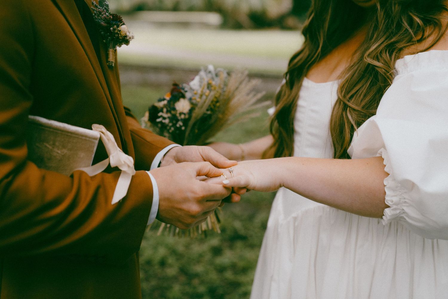 Hands exchanging wedding rings during an outdoor ceremony with a bouquet of lavender flowers in view.
