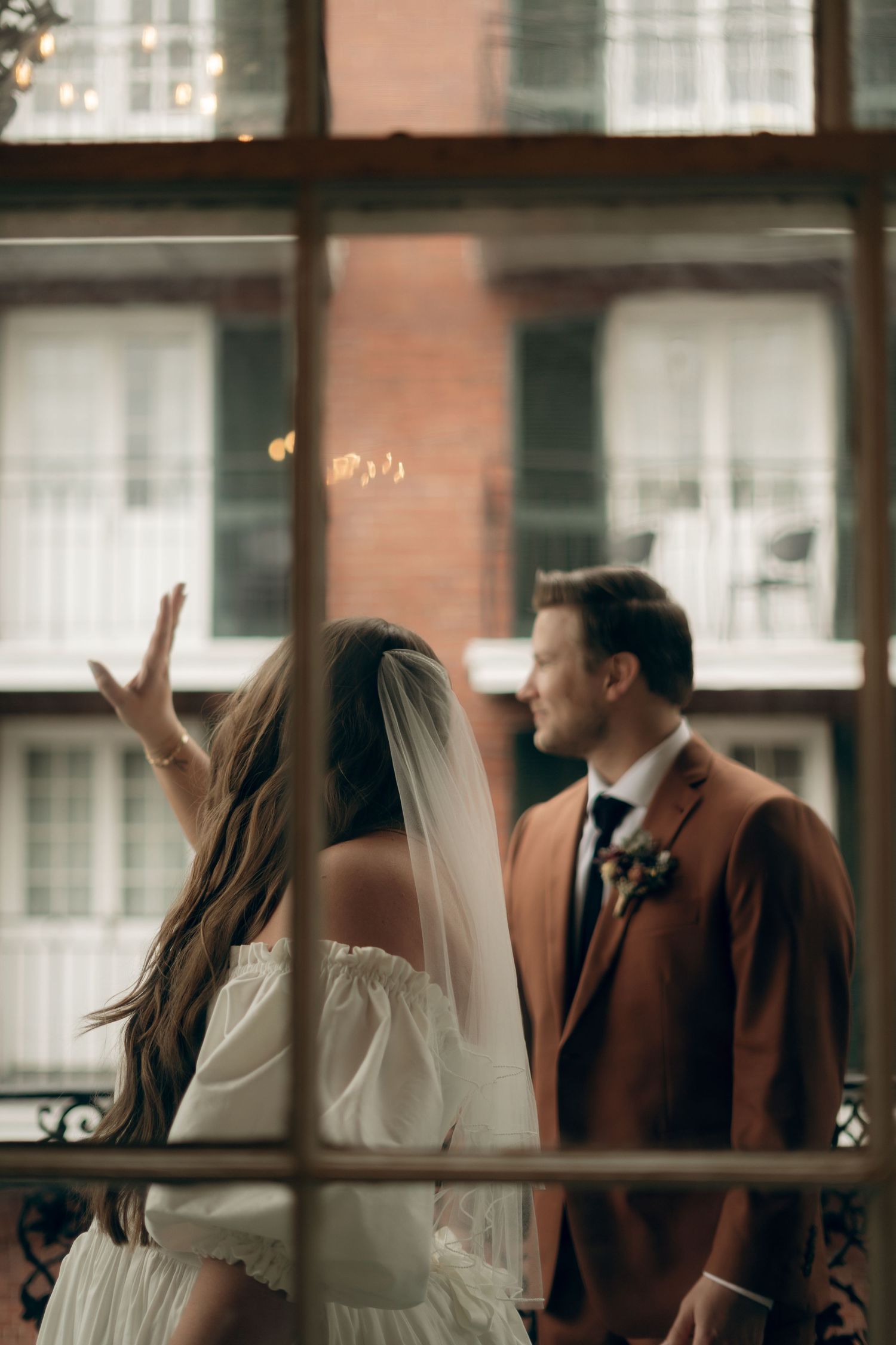 A couple in wedding attire share a moment on a balcony with windows and sun flares in the background.