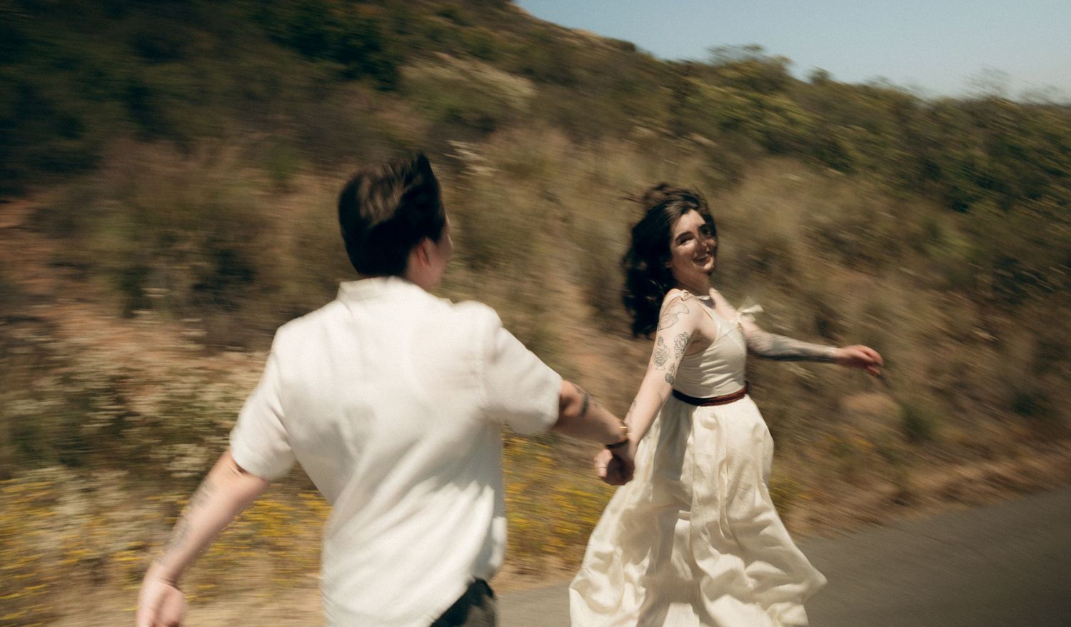A couple runs together on a dusty hillside path, with the person in a flowing white dress twirling joyfully.