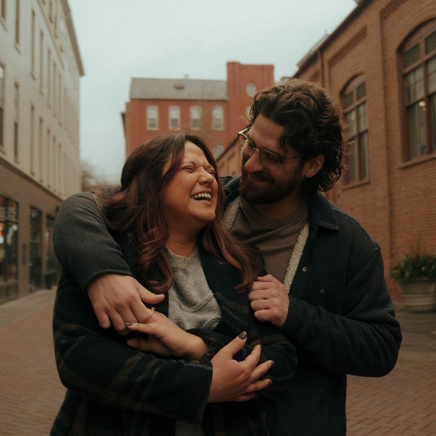 Joyful couple laughing and embracing on brick-paved street with rustic buildings in background.