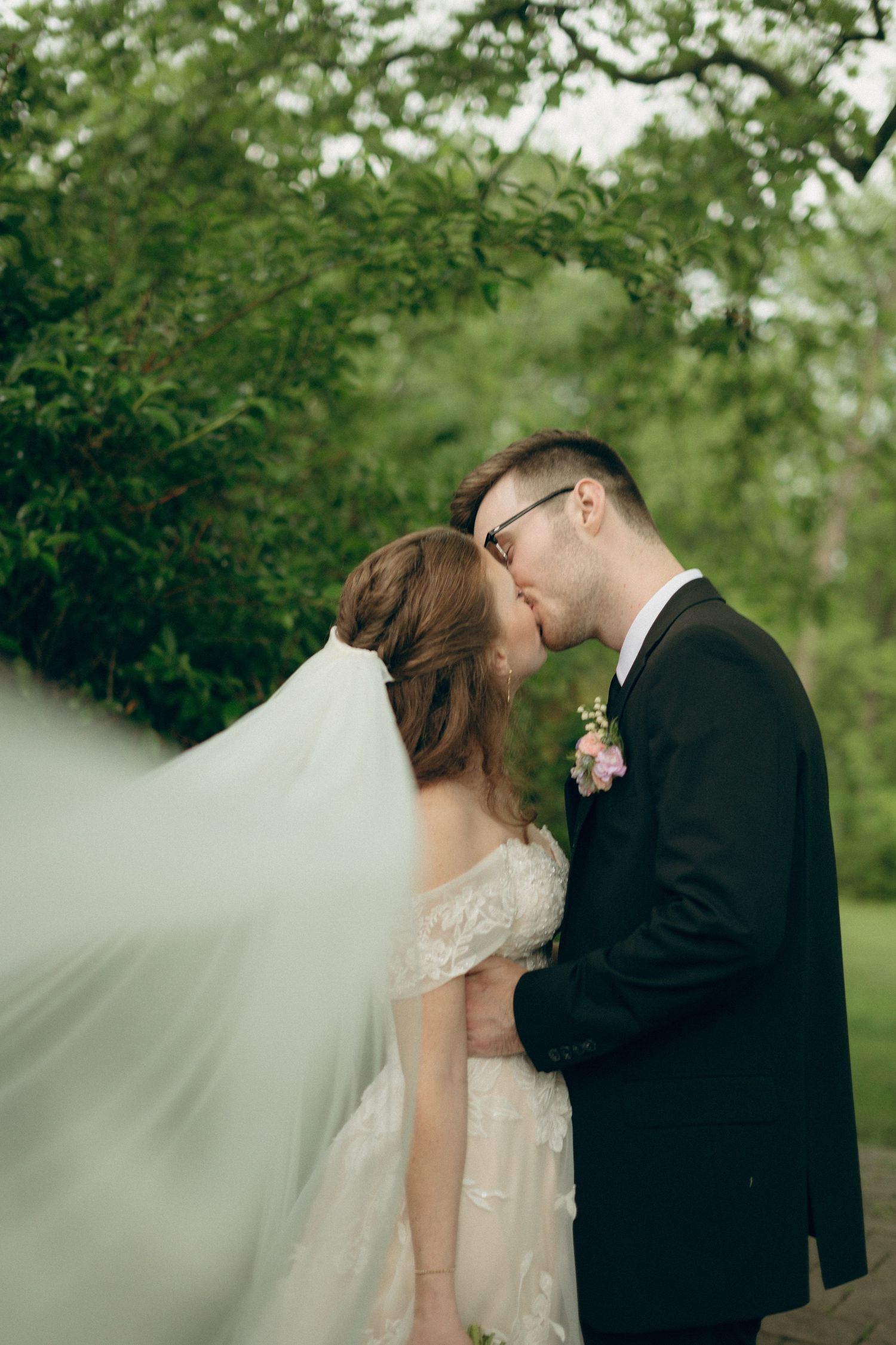 Romantic wedding couple shares an intimate moment under trees with the bride's veil flowing in the breeze.