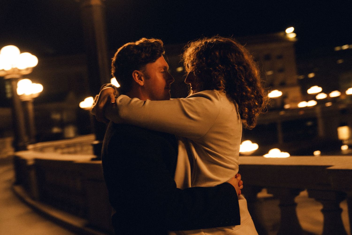 Romantic couple embracing and kissing at night with glowing city lights in the background.