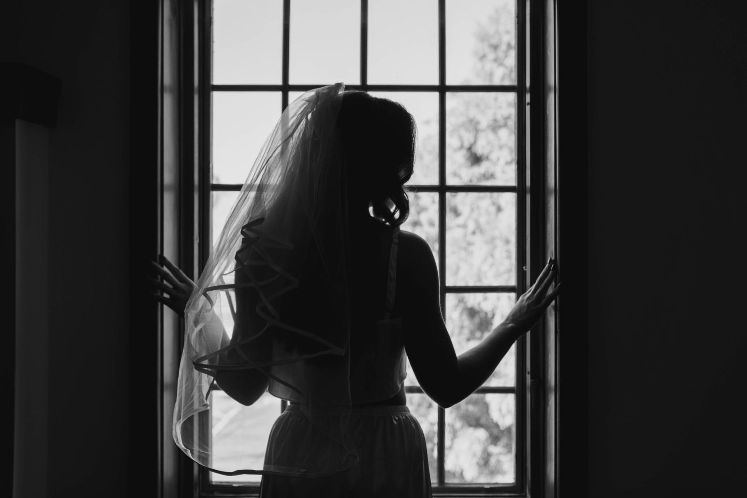 Silhouette of a bride standing by a large window in dramatic black and white lighting.
