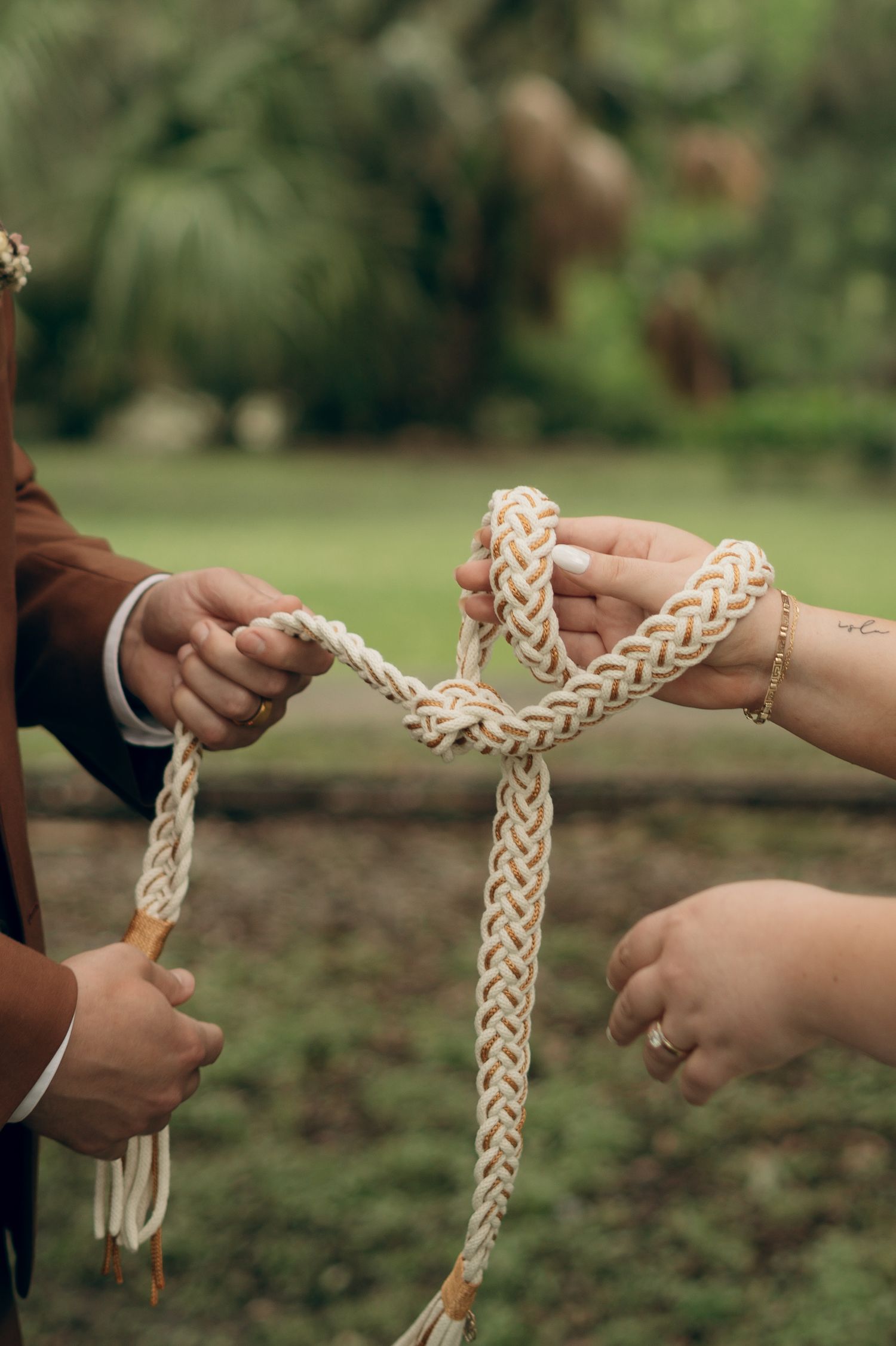 Multiple hands holding and intertwining a braided white ceremonial rope or cord in an outdoor garden setting.