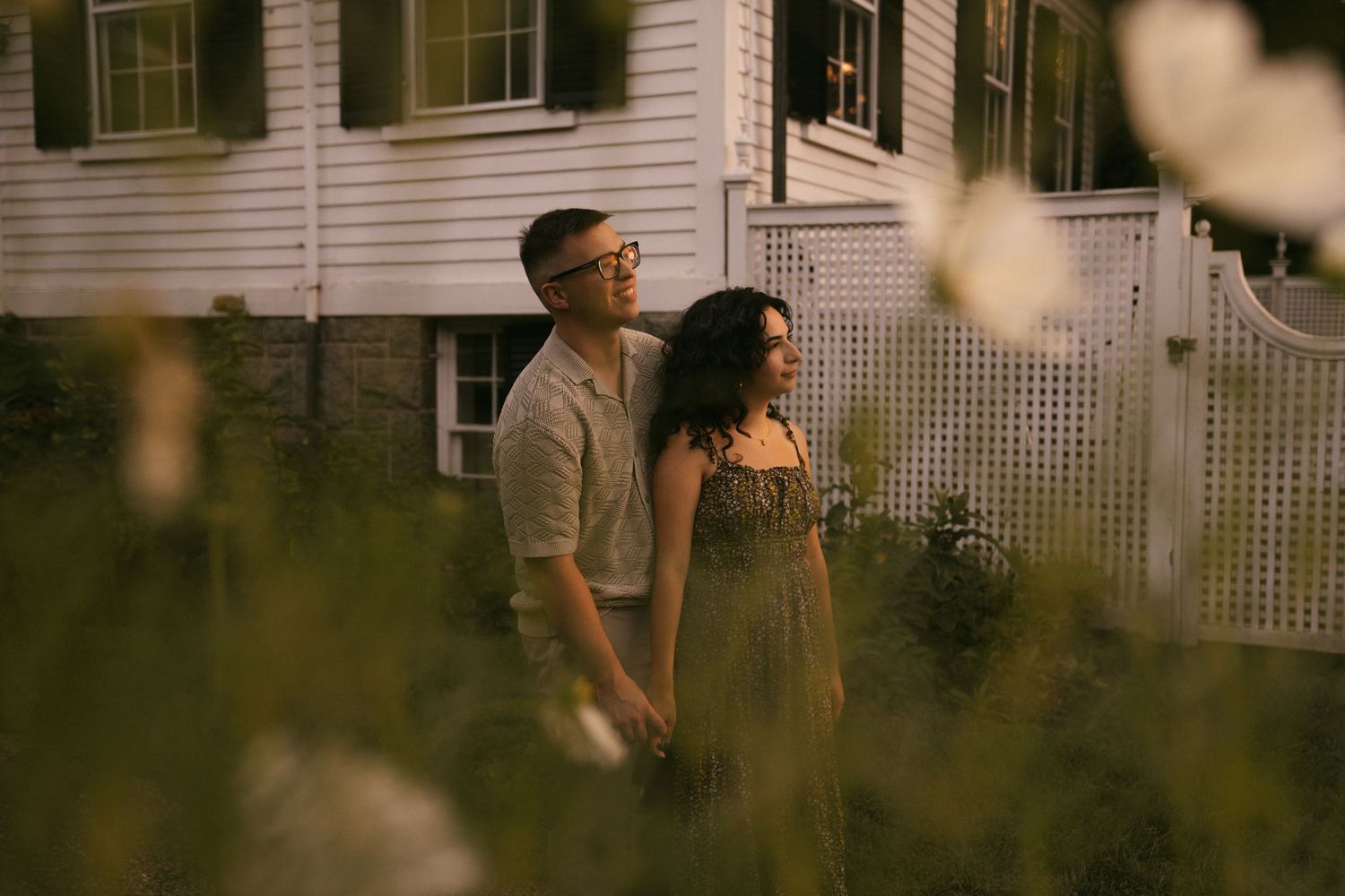 A couple holds hands among wildflowers at dusk, standing before a charming white house with warm glowing windows.