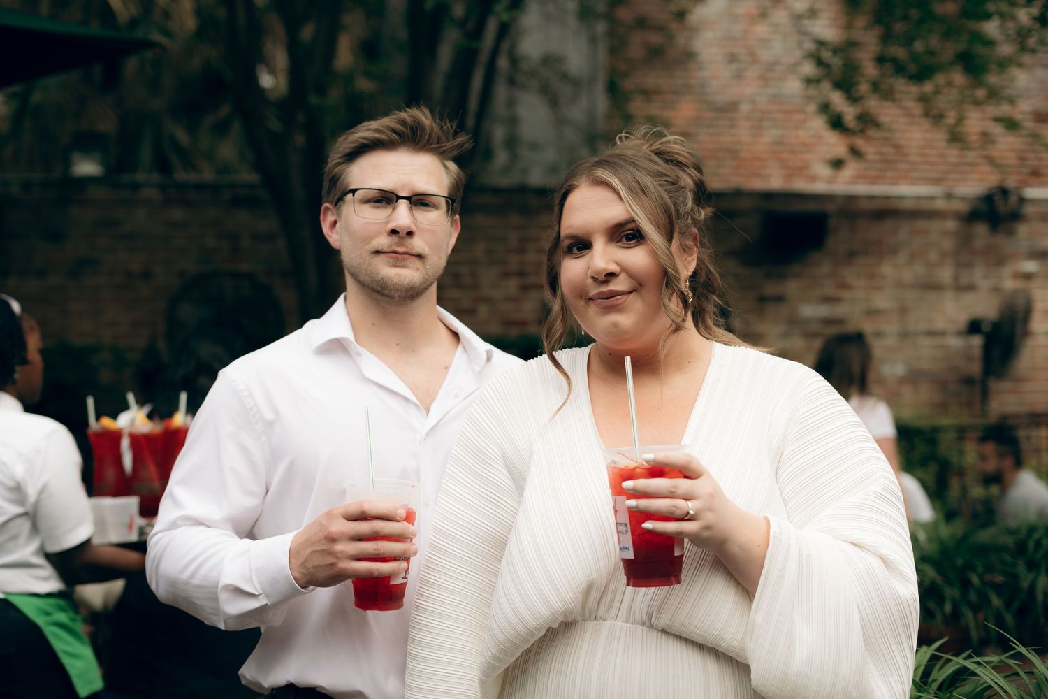Two people in white outfits holding red drinks at a brick wall gathering.