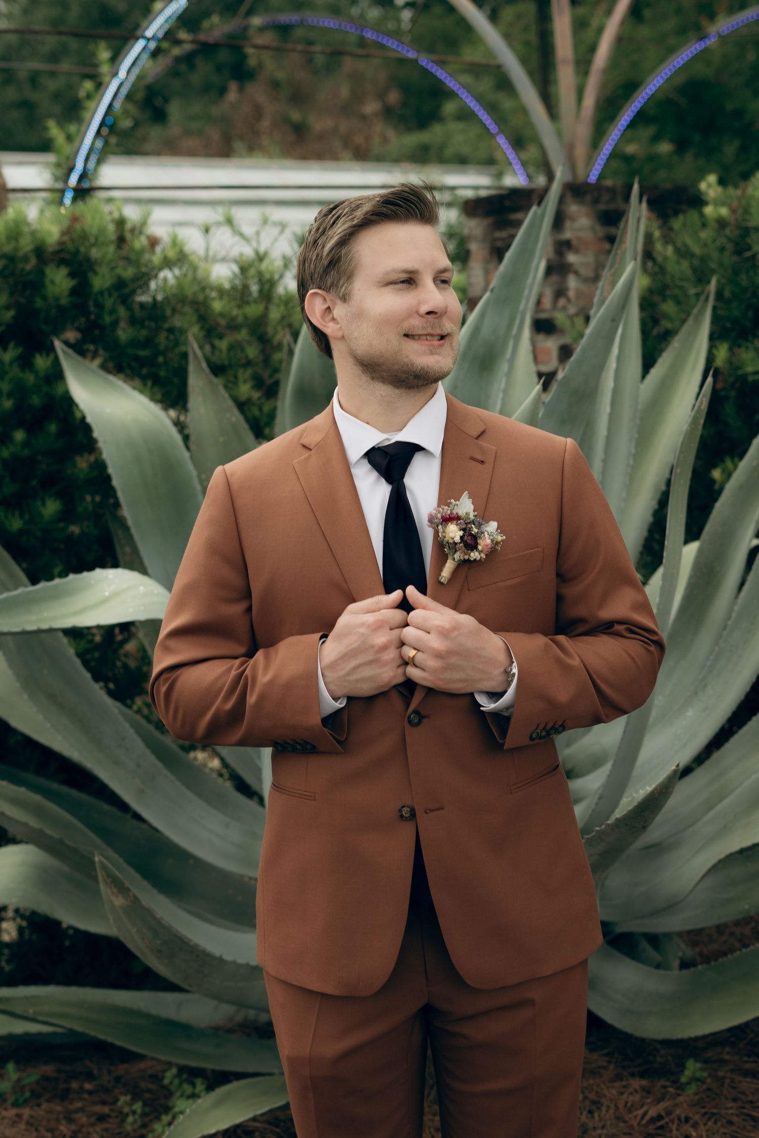 Elegant brown suit and boutonniere photographed in front of large agave plants in garden setting.