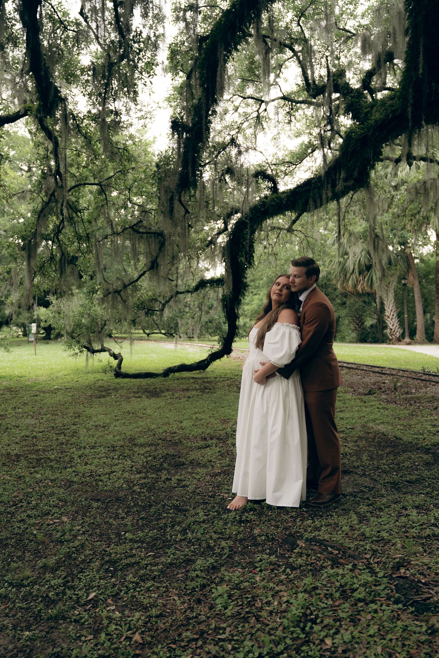 A couple shares an intimate embrace against a backdrop of moss-draped oak trees in a serene woodland setting.