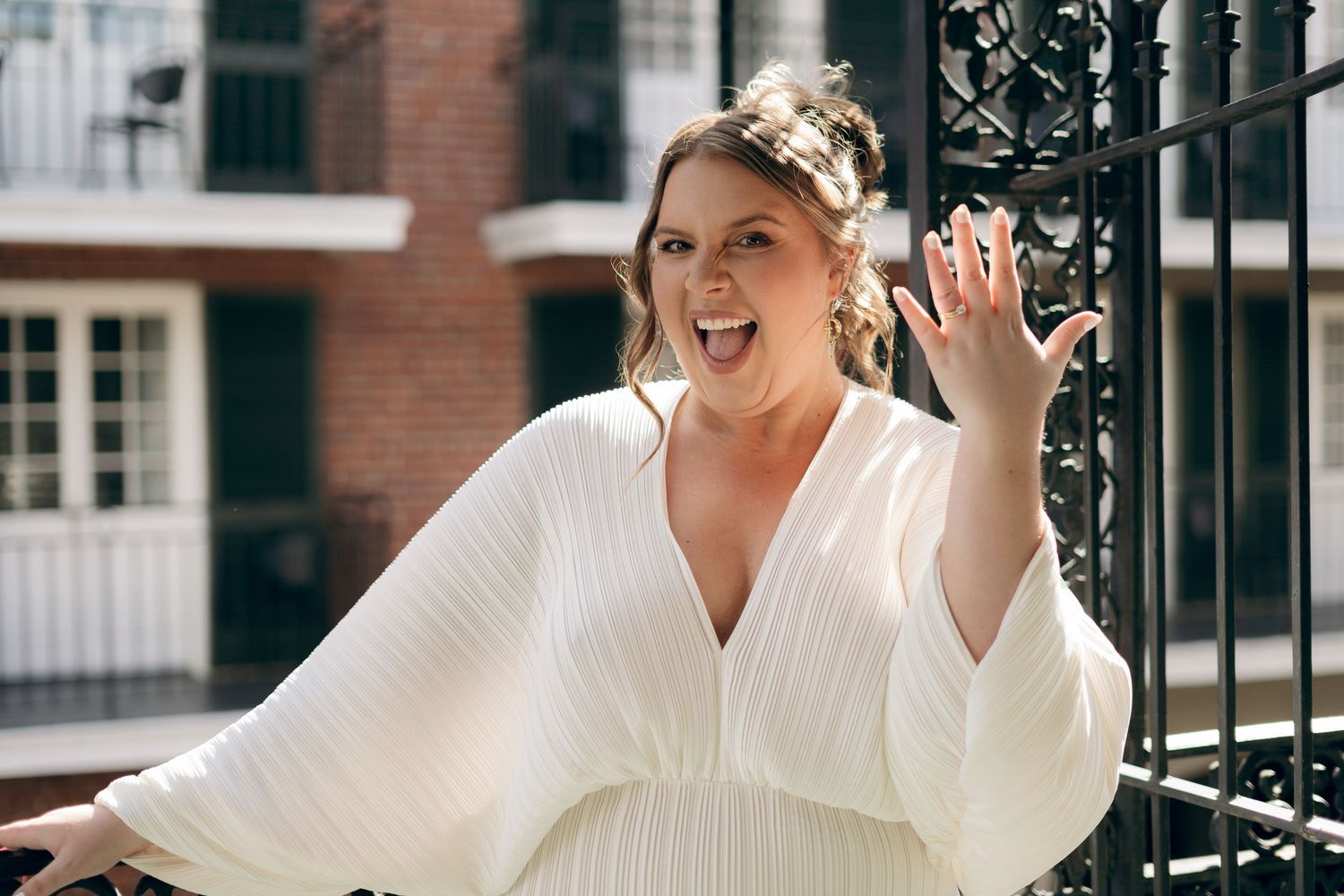 A person in a flowing white v-neck dress with billowing sleeves waves while standing next to a black metal gate.